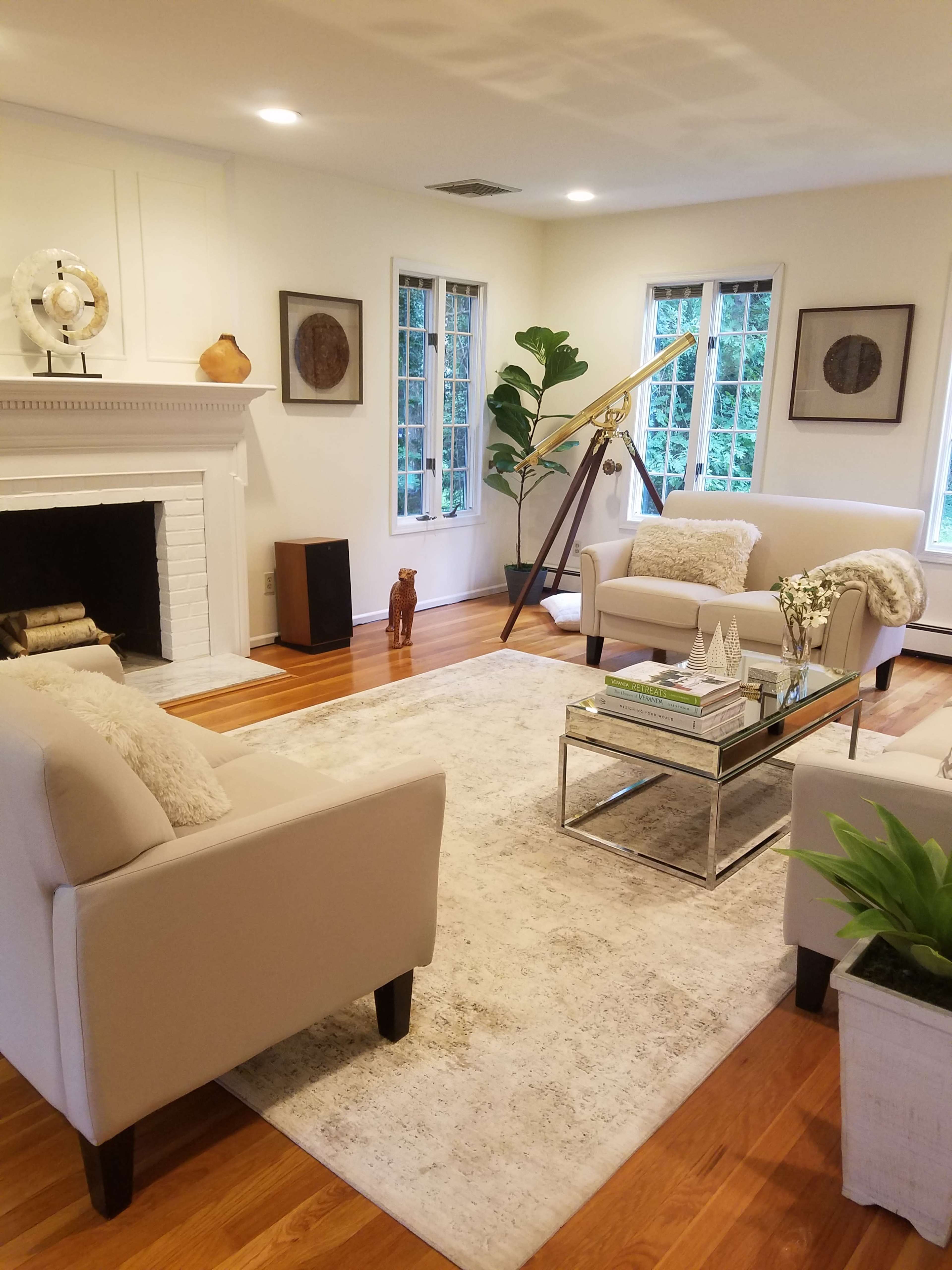 The image shows a living room with two light-colored armchairs, a glass coffee table, a decorative rug, and large windows, featuring a modern fireplace and some indoor plants.