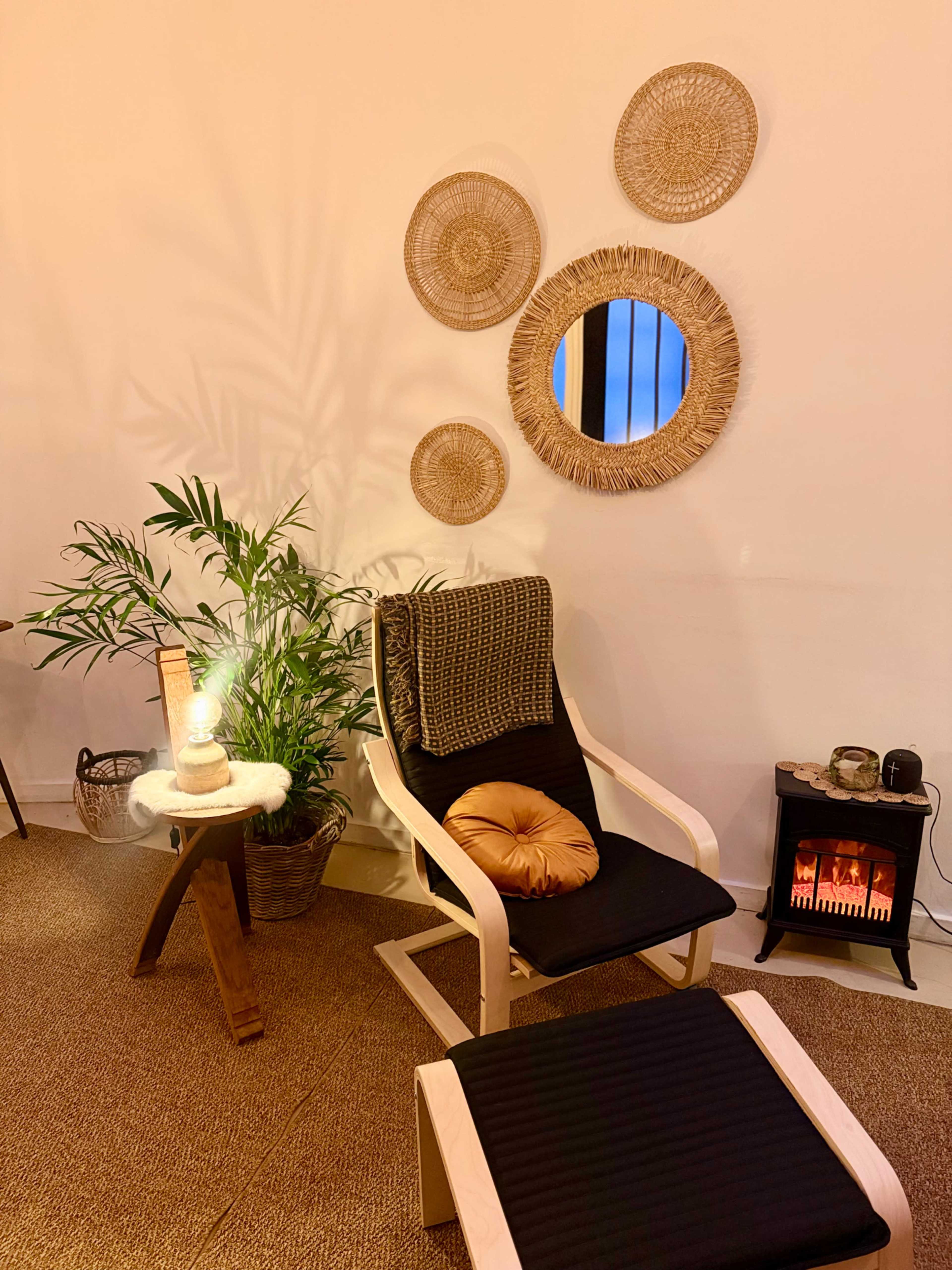 A cozy indoor space features a comfortable chair with a footrest, a small side table with a glowing candle, and decorative wall plates next to a plant and a small electric heater emitting warmth.