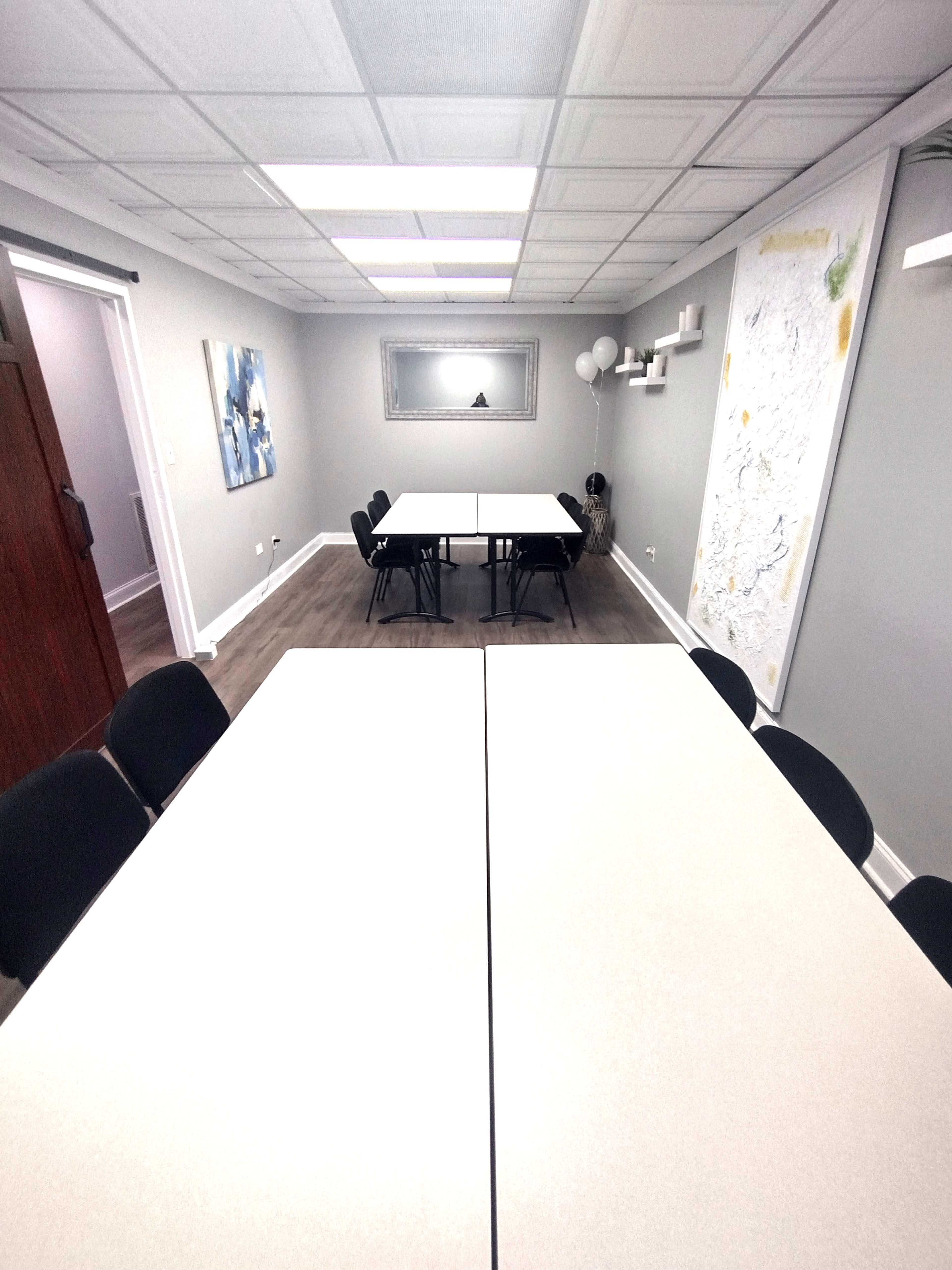 A meeting room with two rectangular tables arranged in the center and black chairs surrounding them, along with a whiteboard and artwork on the walls.