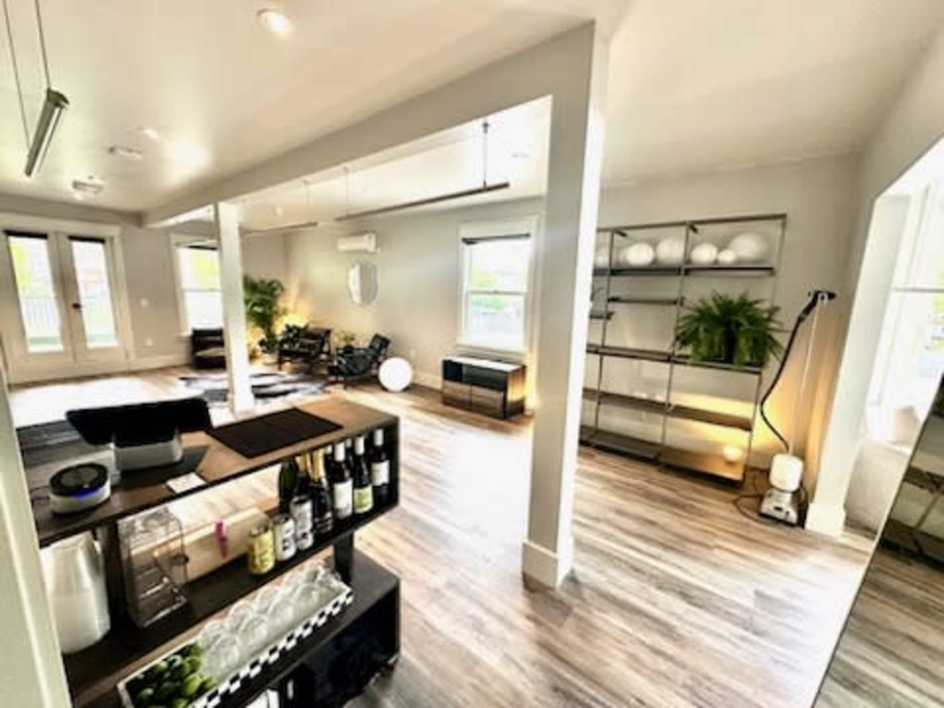 A modern, open-concept living space with wood flooring, a bar cart, shelves with decorative items, and large windows providing natural light.