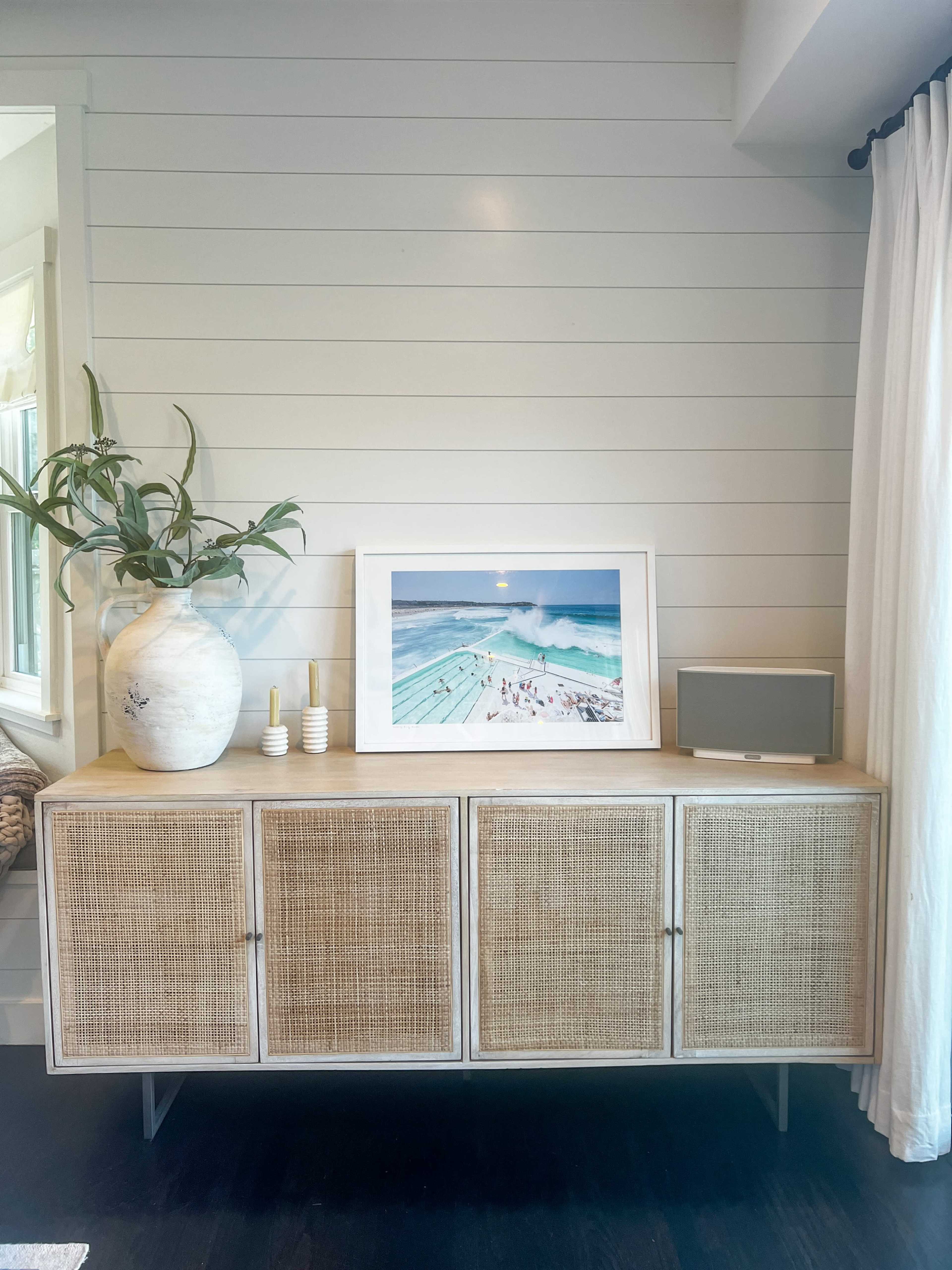 A light-colored cabinet with woven doors holds a framed beach photograph and decorative items, beside a potted plant and a tablet.