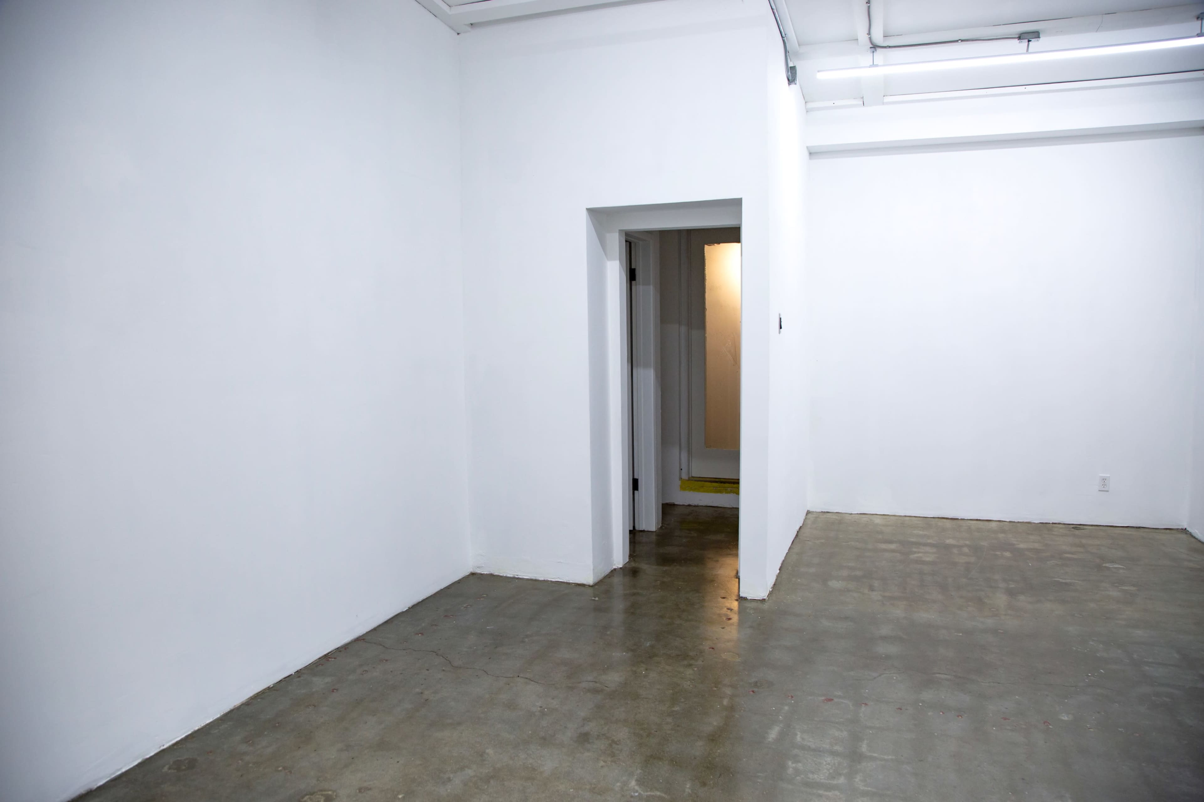 The image shows a minimalist room with white walls and a concrete floor, featuring an open doorway leading to a hallway.