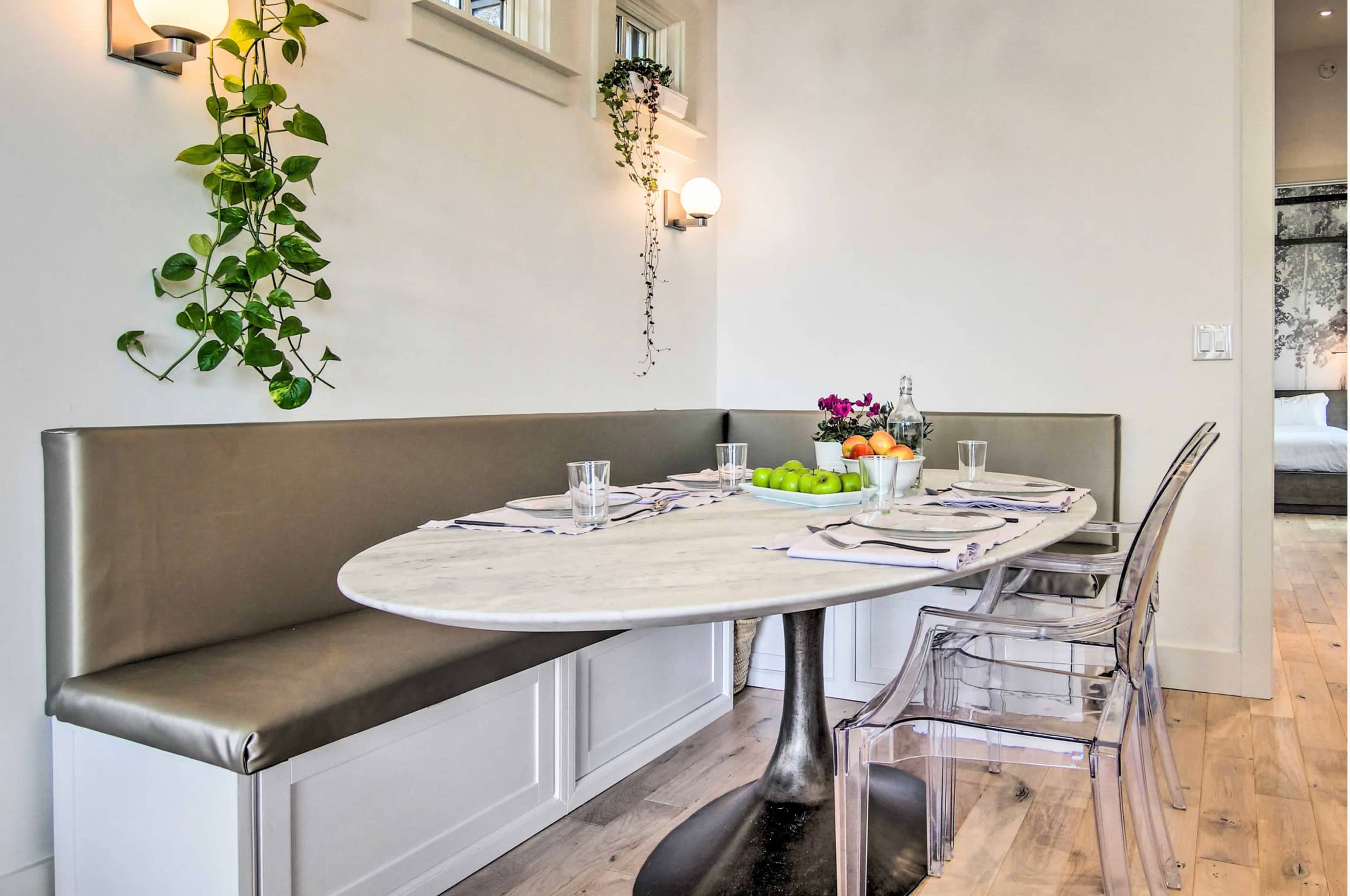 A round marble table is set with fruit and glassware in a bright dining area featuring a built-in upholstered bench and clear acrylic chairs.
