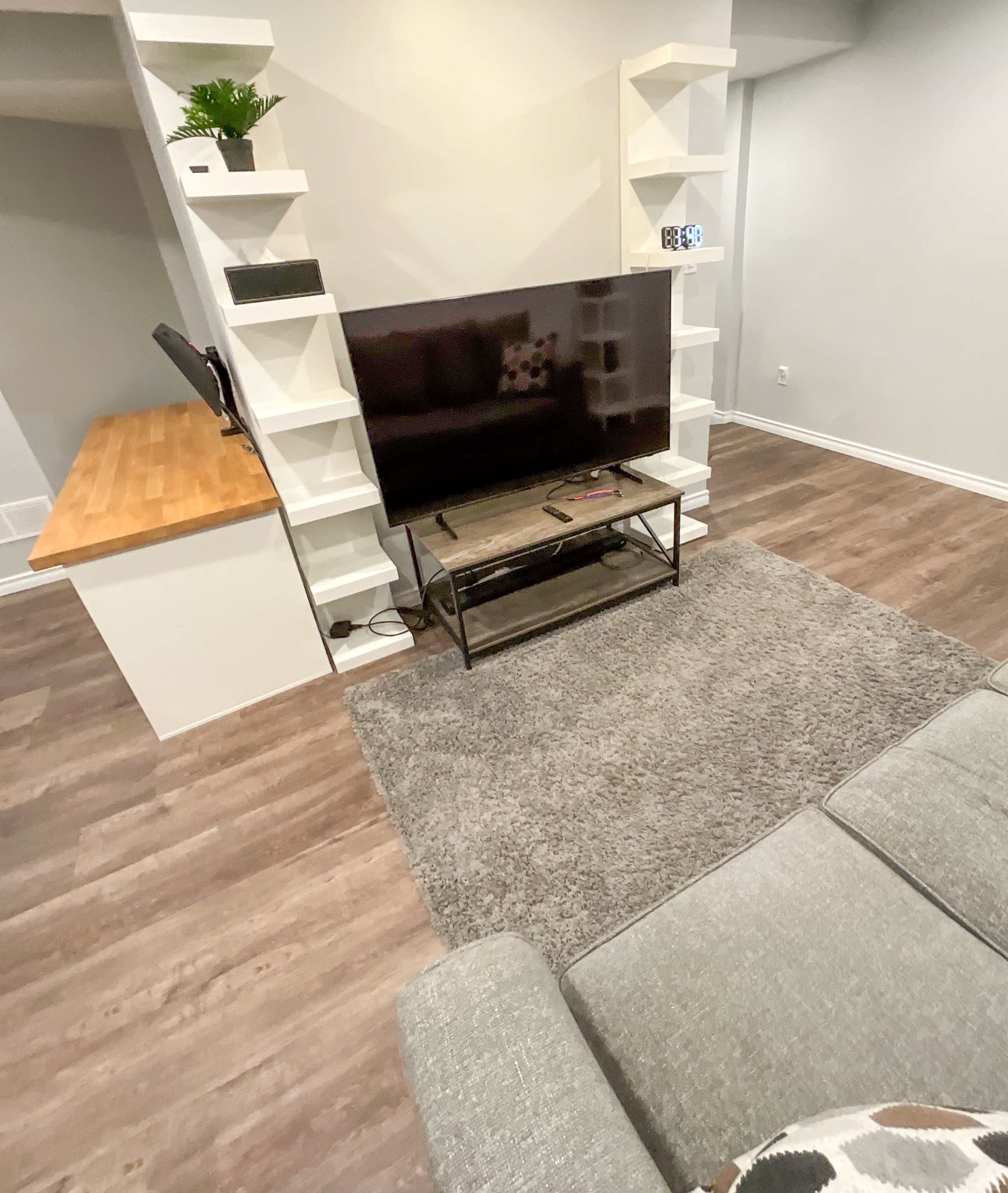 The image shows a cozy living space featuring a flat-screen TV on a stand, a gray couch, a light wood bar area, and gray carpeting, with white shelves flanking the TV.