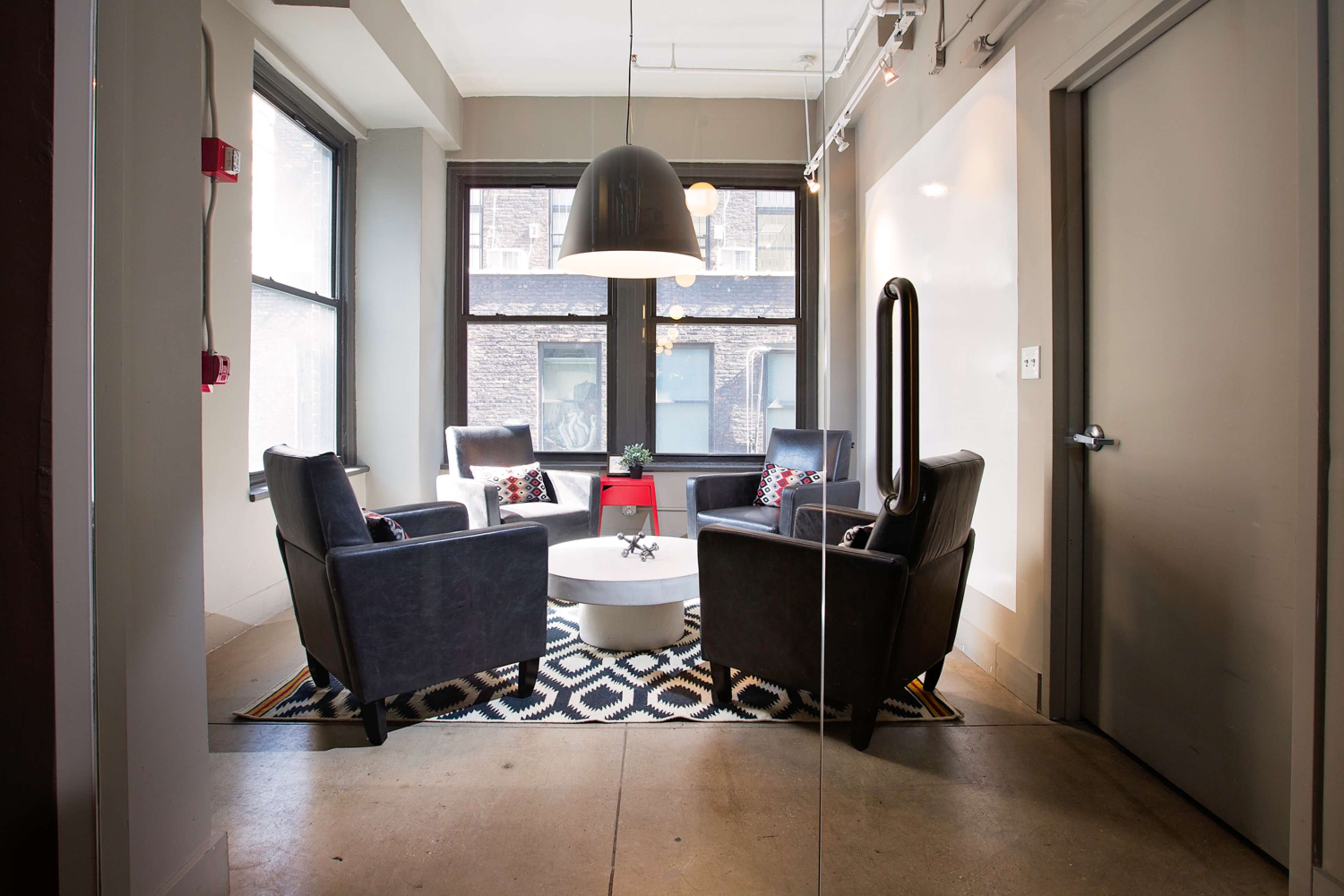 The image shows a small meeting area with four chairs arranged around a circular table, under a large pendant light, with large windows letting in natural light.