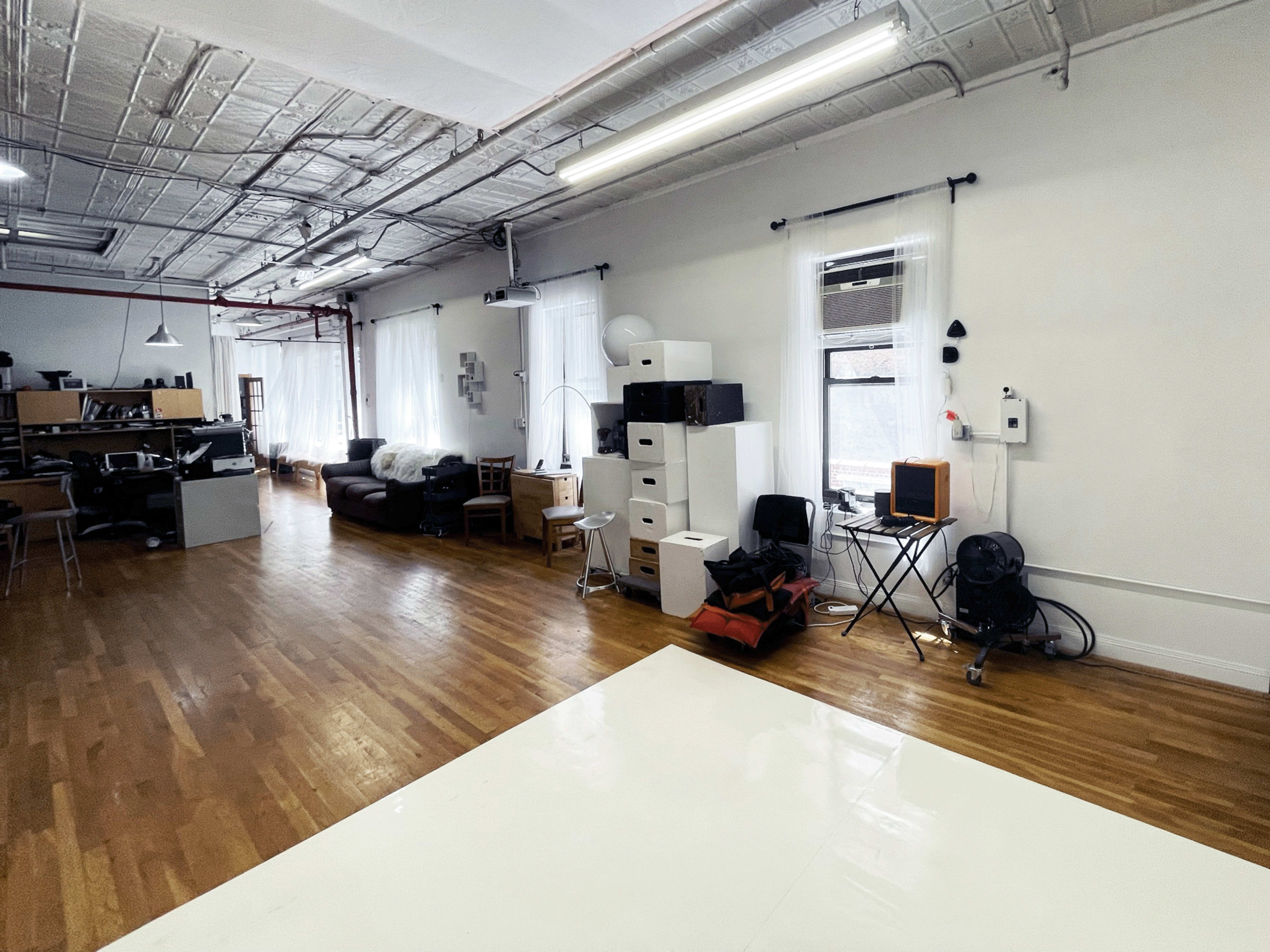 The image shows a spacious, well-lit studio or office space with wooden flooring, furniture including a couch and desk, and various storage units scattered throughout.