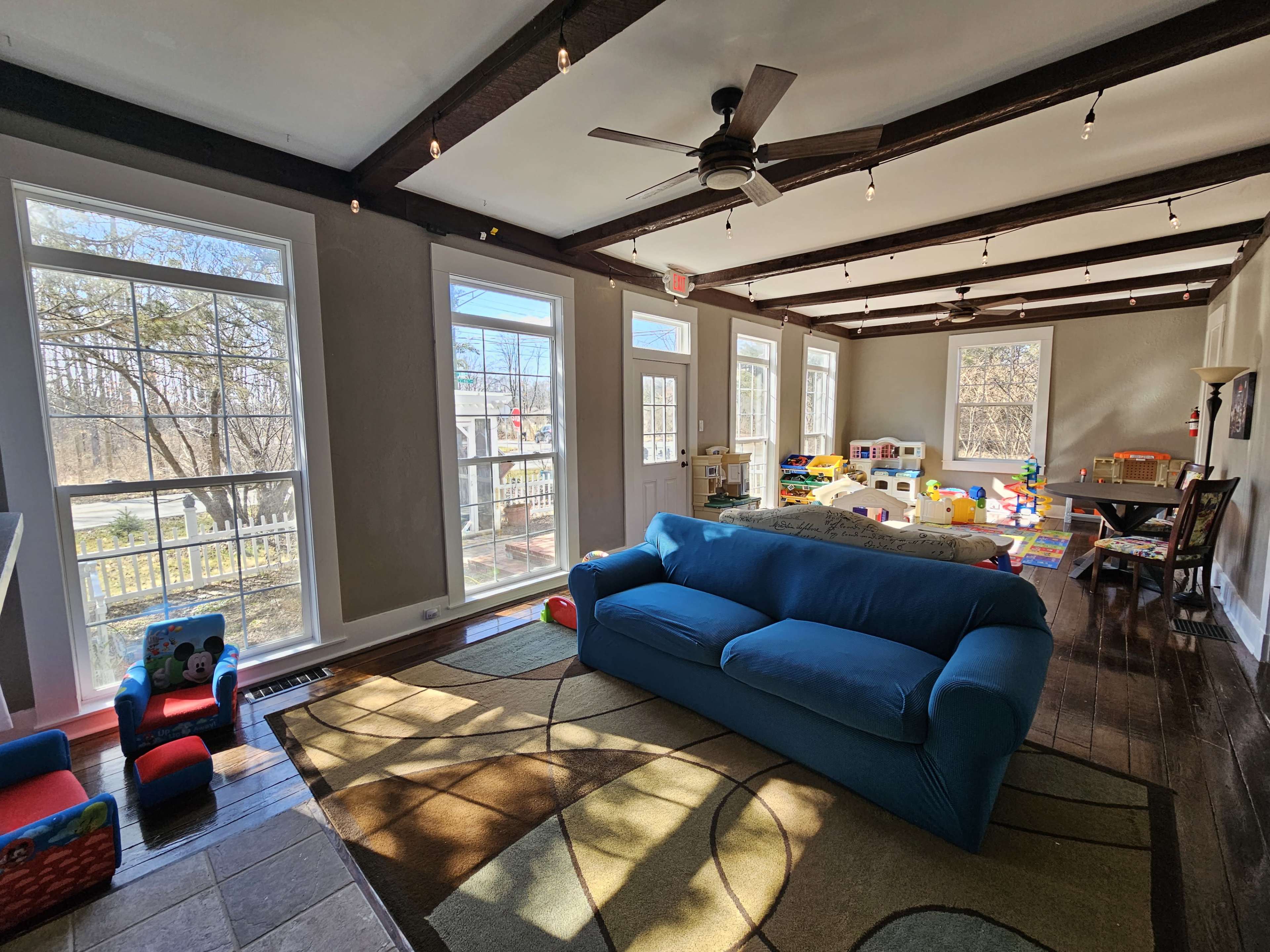 A bright, spacious room features a blue sofa, large windows, and a play area with colorful toys.