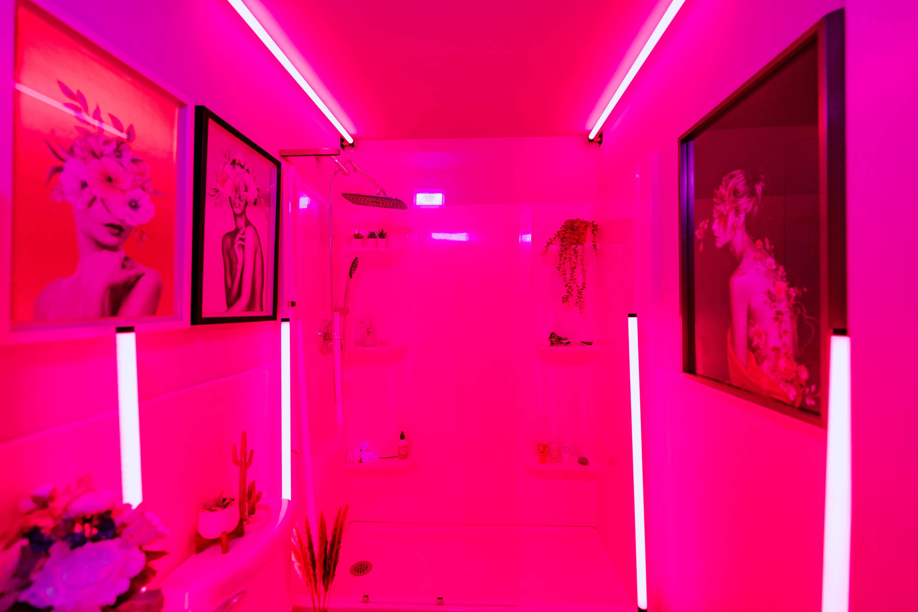 The image shows a shower room with bright pink lighting and two framed artworks on the walls.