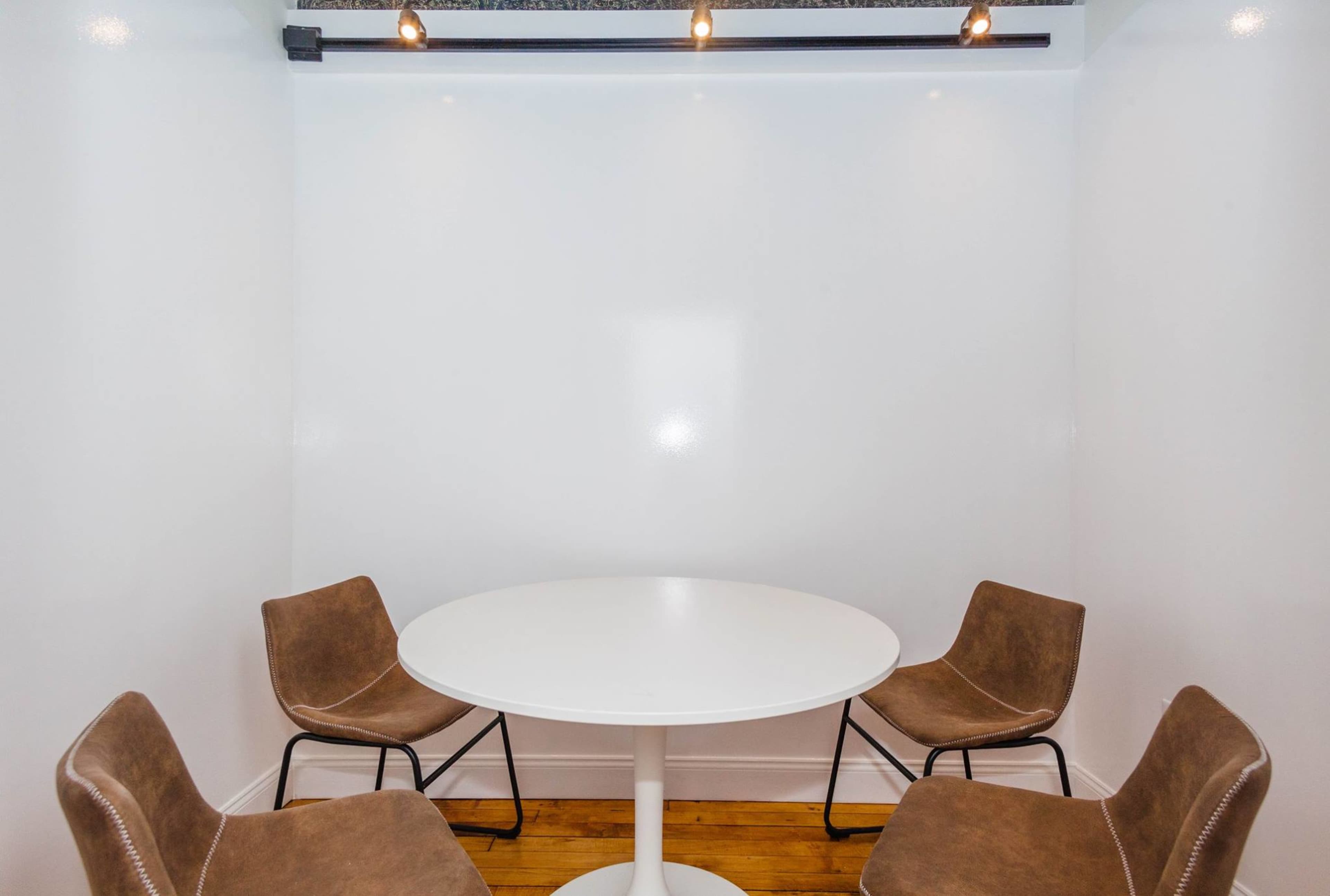 A small, empty room features a white circular table surrounded by four brown chairs.