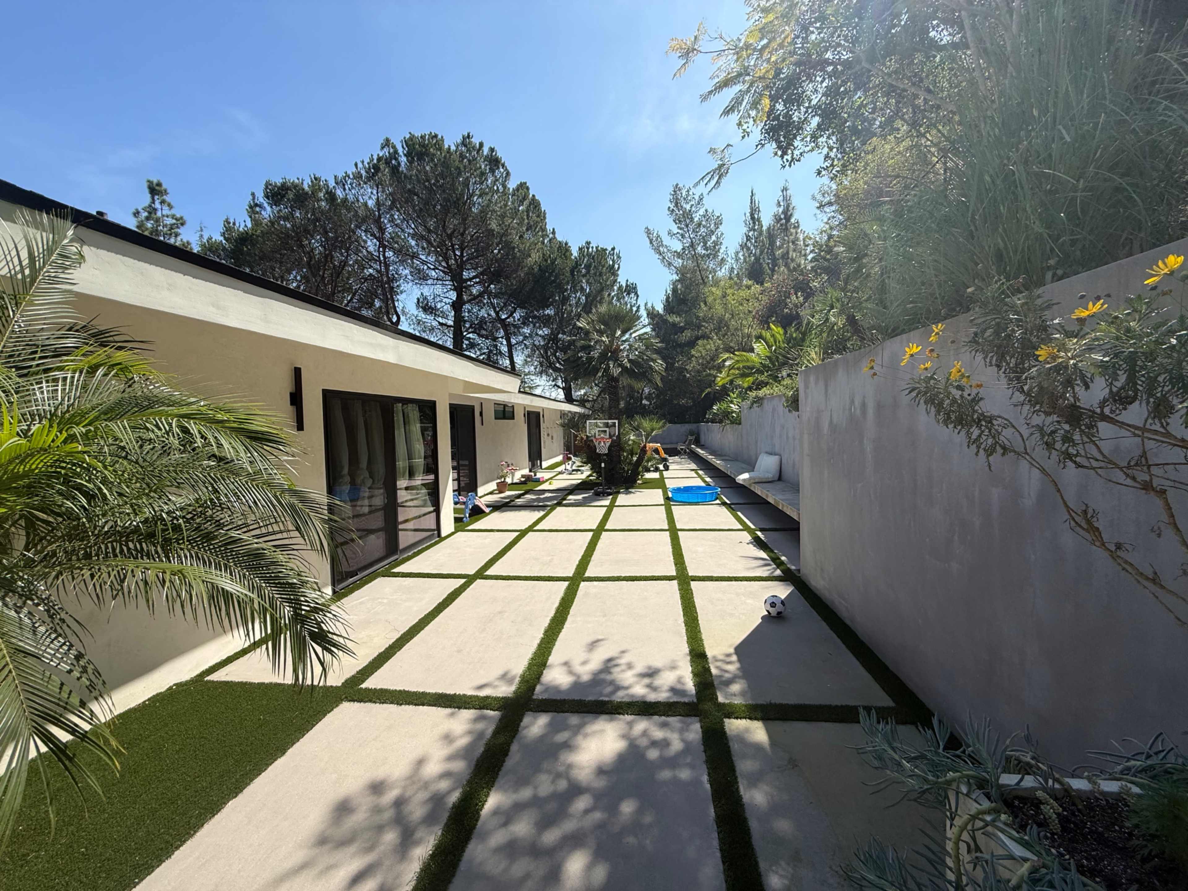 The image shows a modern outdoor area with a decorative pathway lined by greenery and a pool in the background.