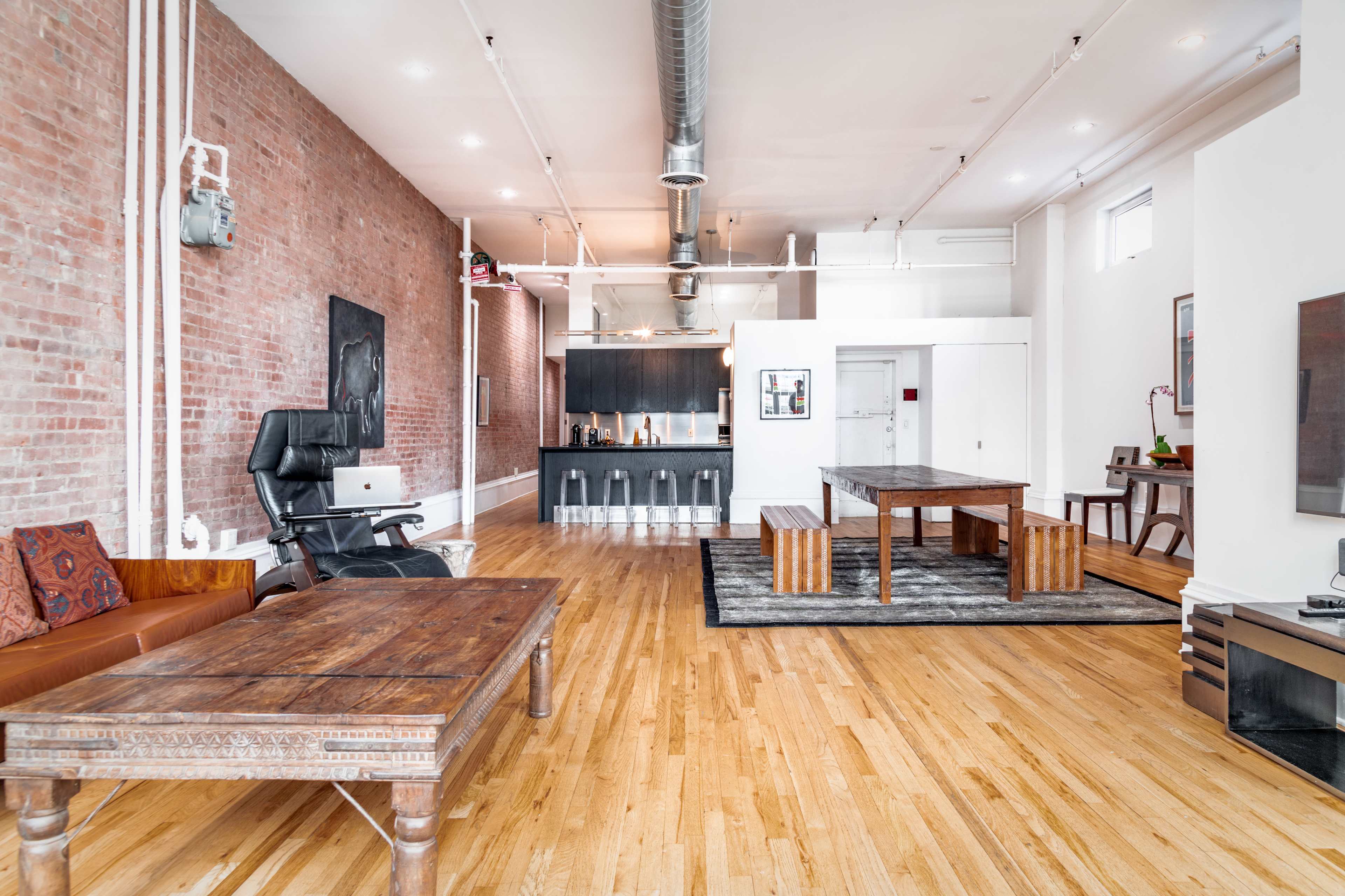 A spacious interior featuring wooden flooring, exposed brick walls, a large dining table, a lounge area with a dark chair, and a kitchen area in the background.