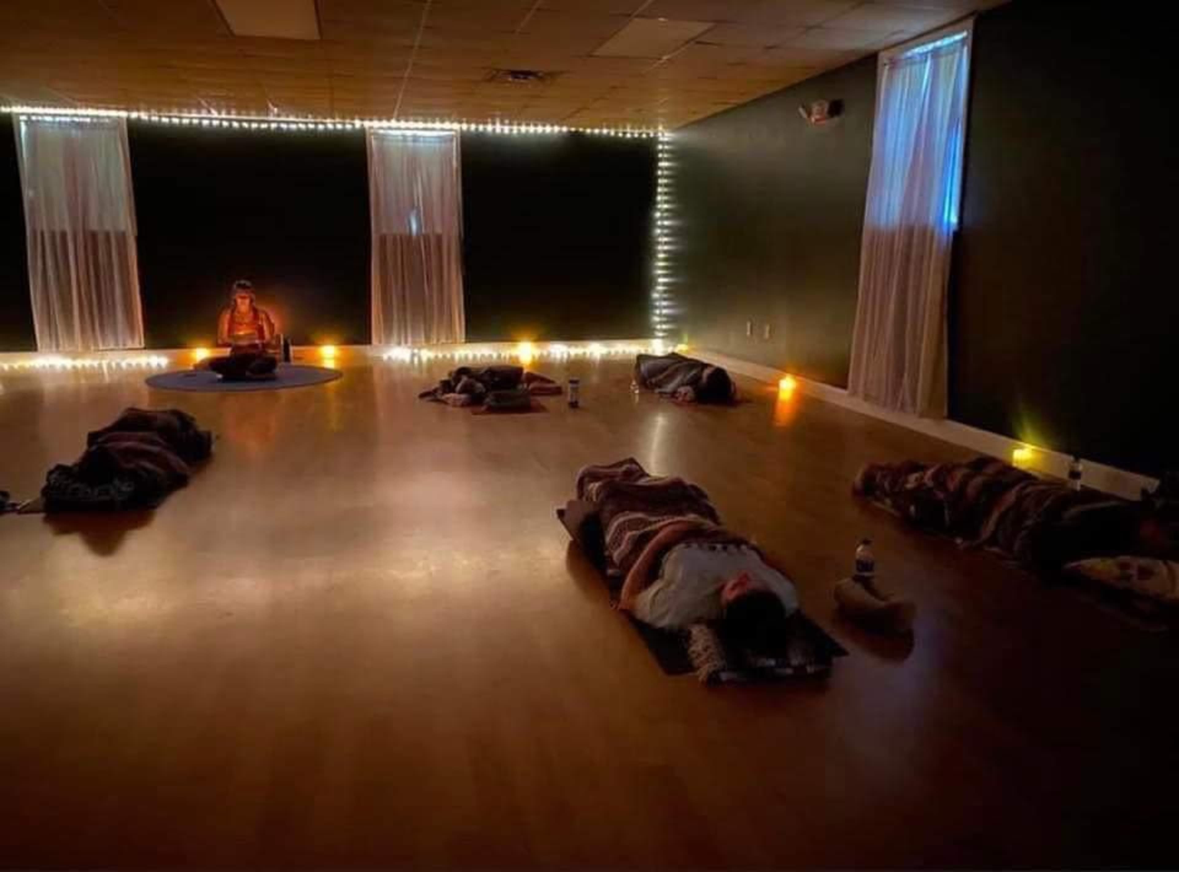 A dimly lit yoga studio features a group of participants lying on mats with blankets, while a seated instructor sits in front, surrounded by candles and string lights.