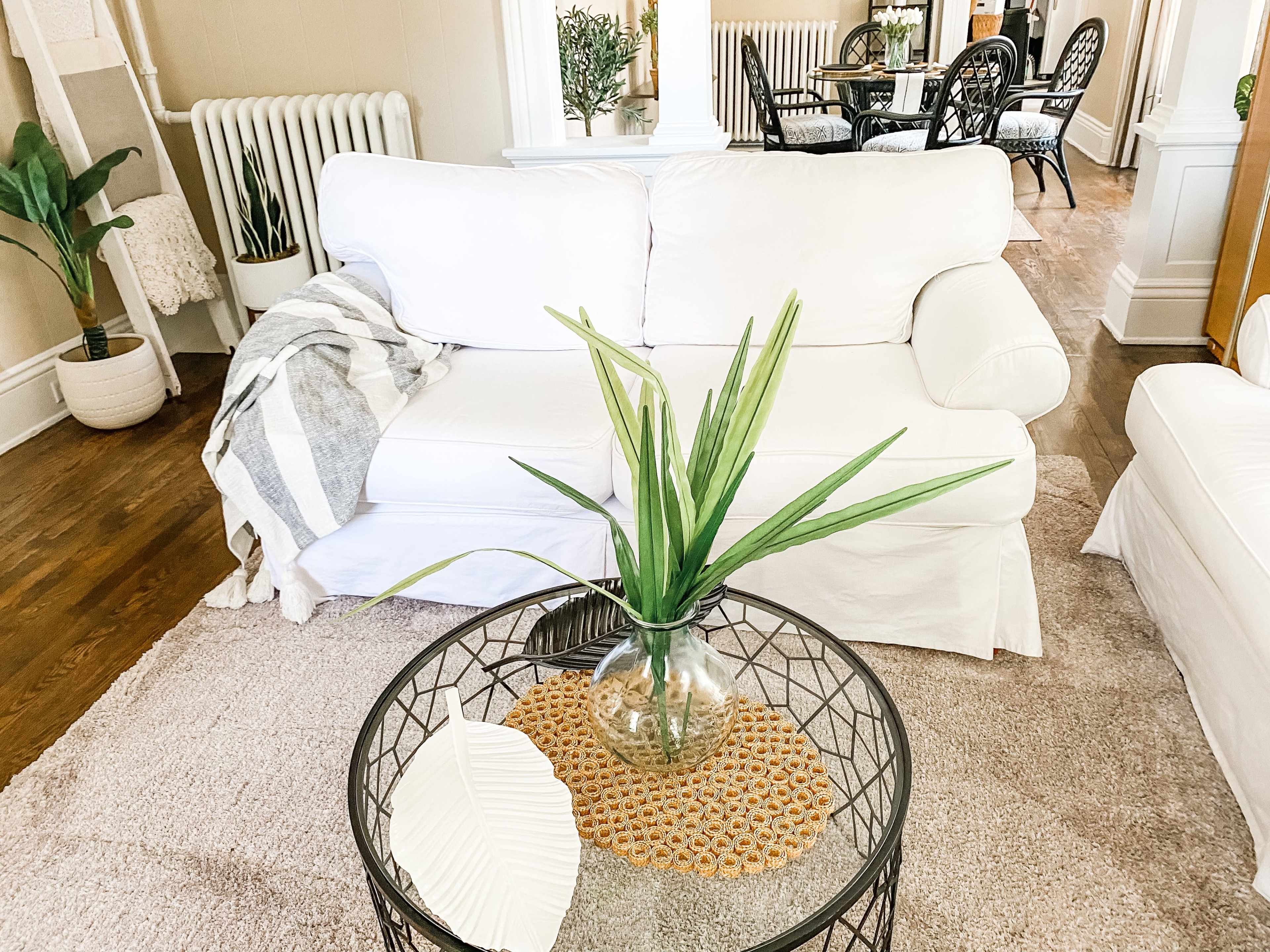 A white sofa with a decorative throw blanket sits next to a round coffee table featuring a glass vase filled with a plant and a decorative leaf.
