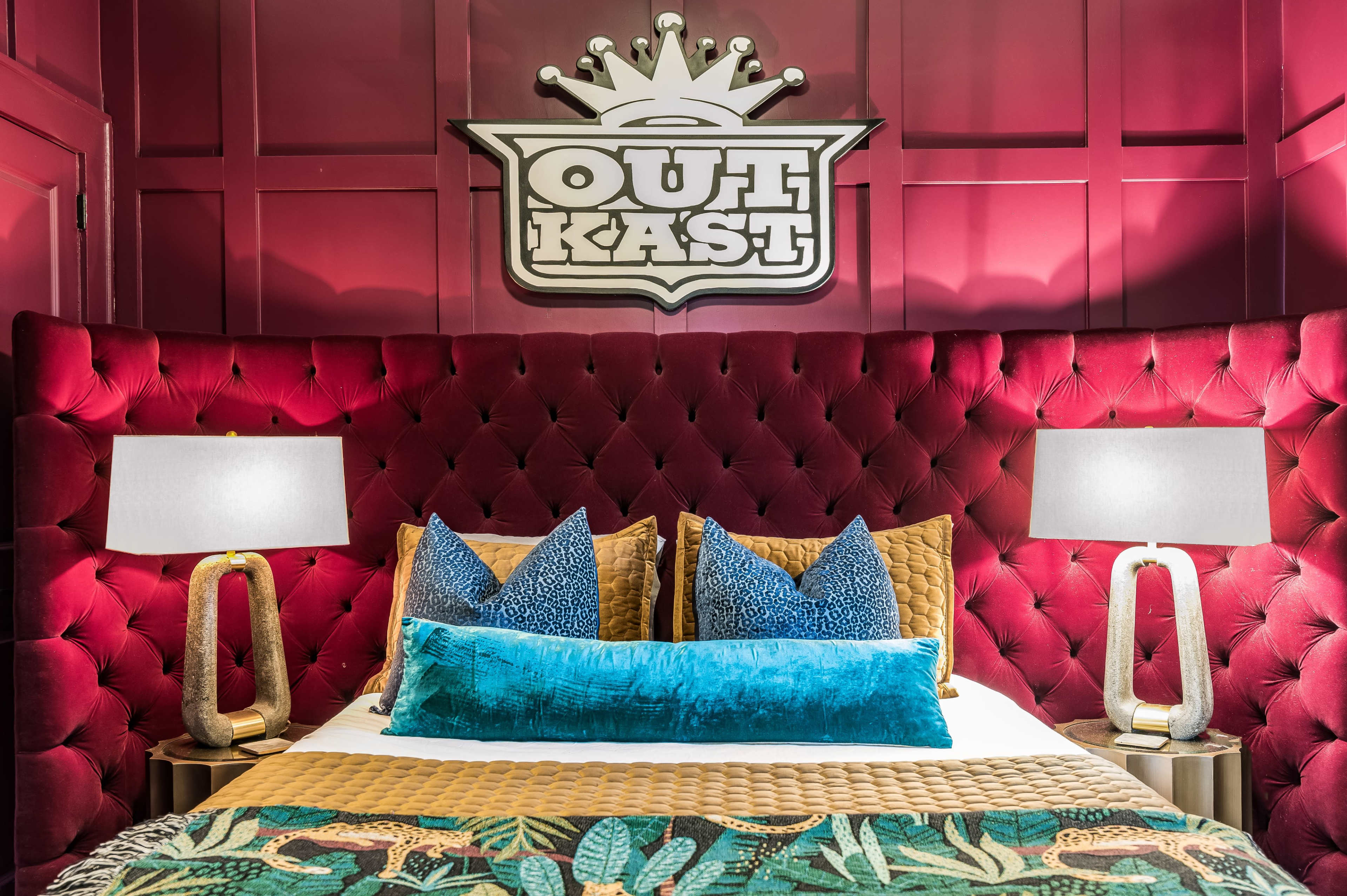 The image shows a stylized hotel room with a plush red upholstered headboard featuring the "OutKast" logo, flanked by modern table lamps and decorative pillows on the bed.