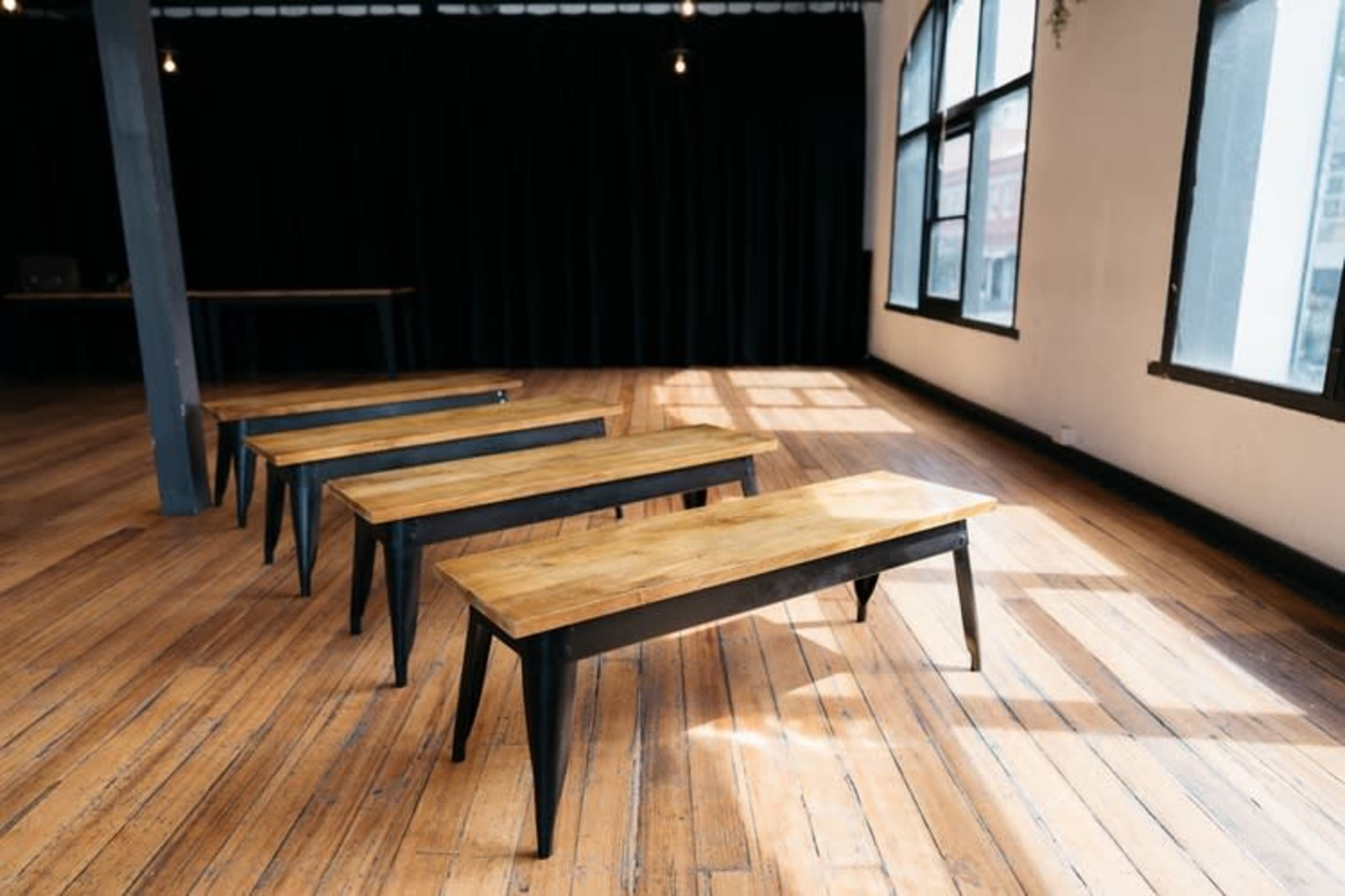 Four wooden benches with metal legs are arranged in a room with wooden flooring and large windows allowing natural light to enter.