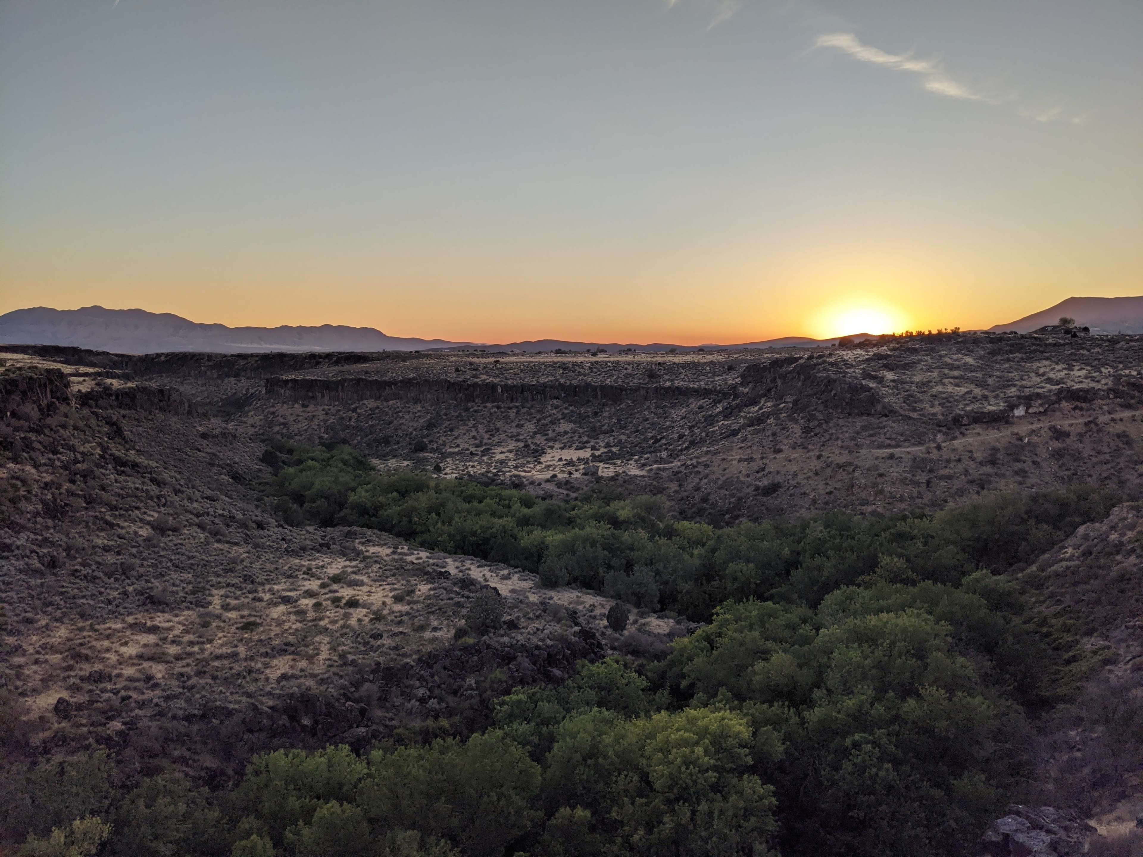 The sun sets over a valley surrounded by rocky cliffs and greenery.