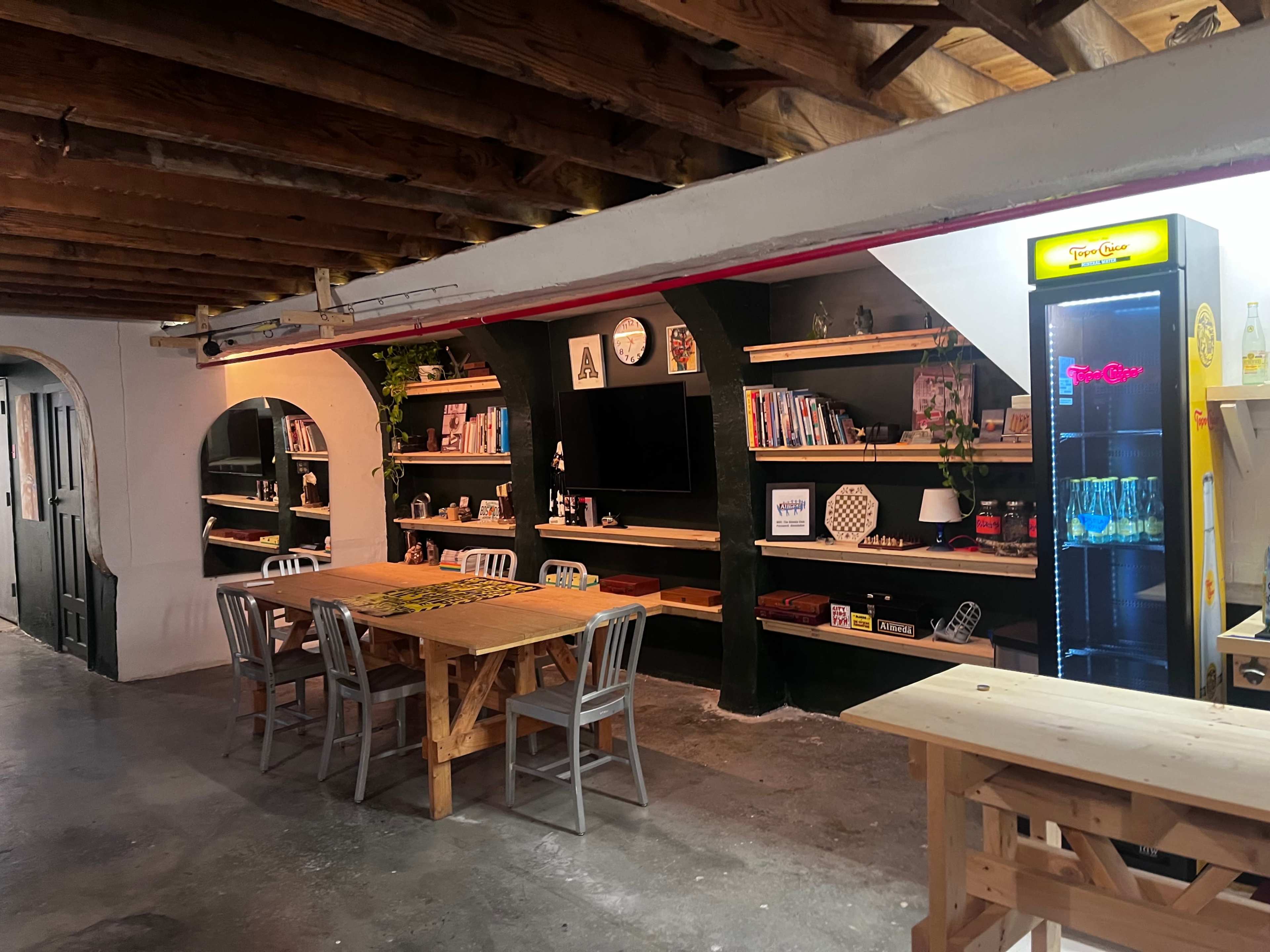 The image shows a spacious room with wooden beams, equipped with a large table surrounded by chairs, shelves filled with various items, and a vending machine in the corner.