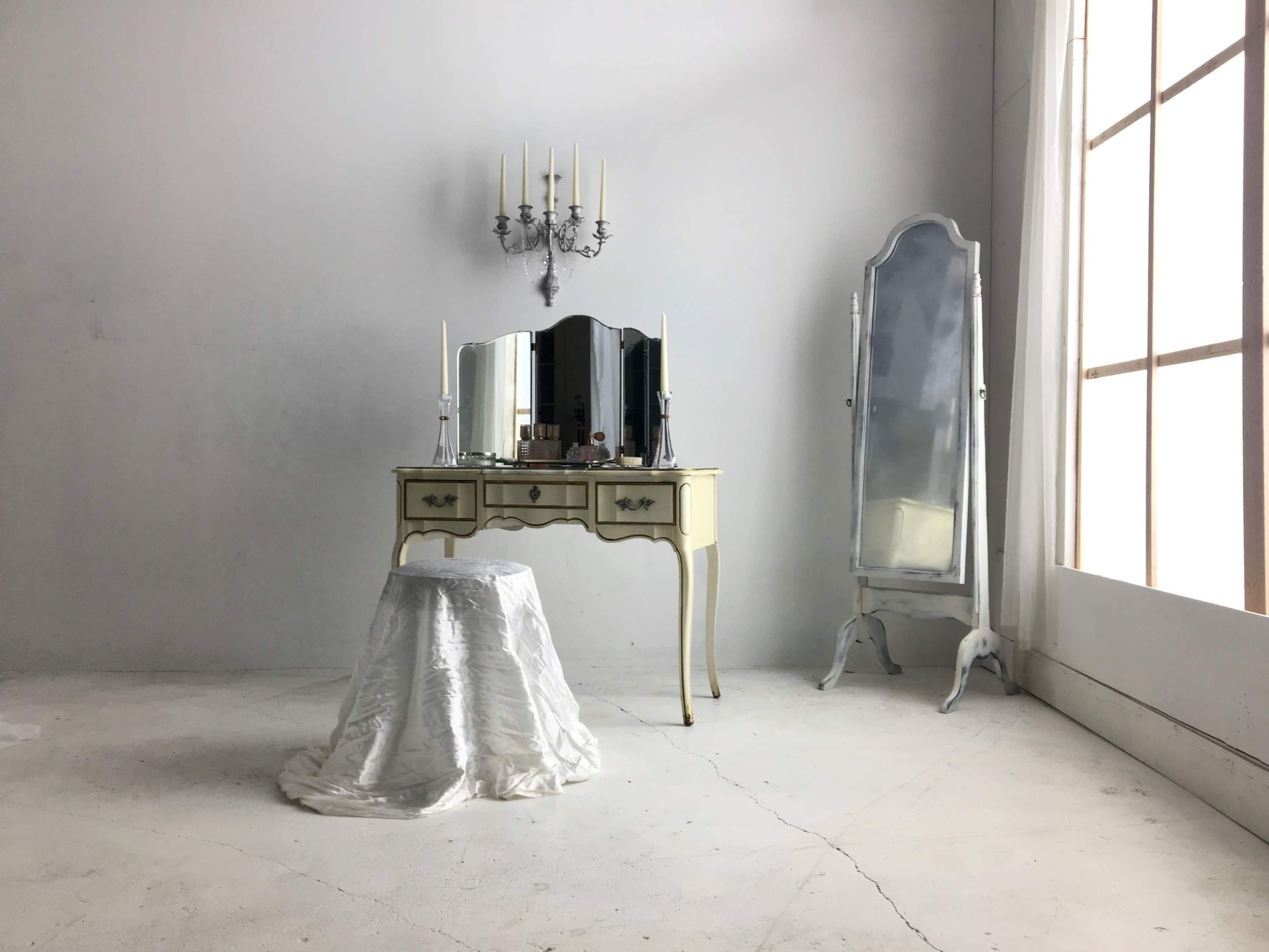 The image shows a minimalist room featuring a vintage vanity table with a mirror, a stool covered in white fabric, and a tall, ornate mirror beside a large window.