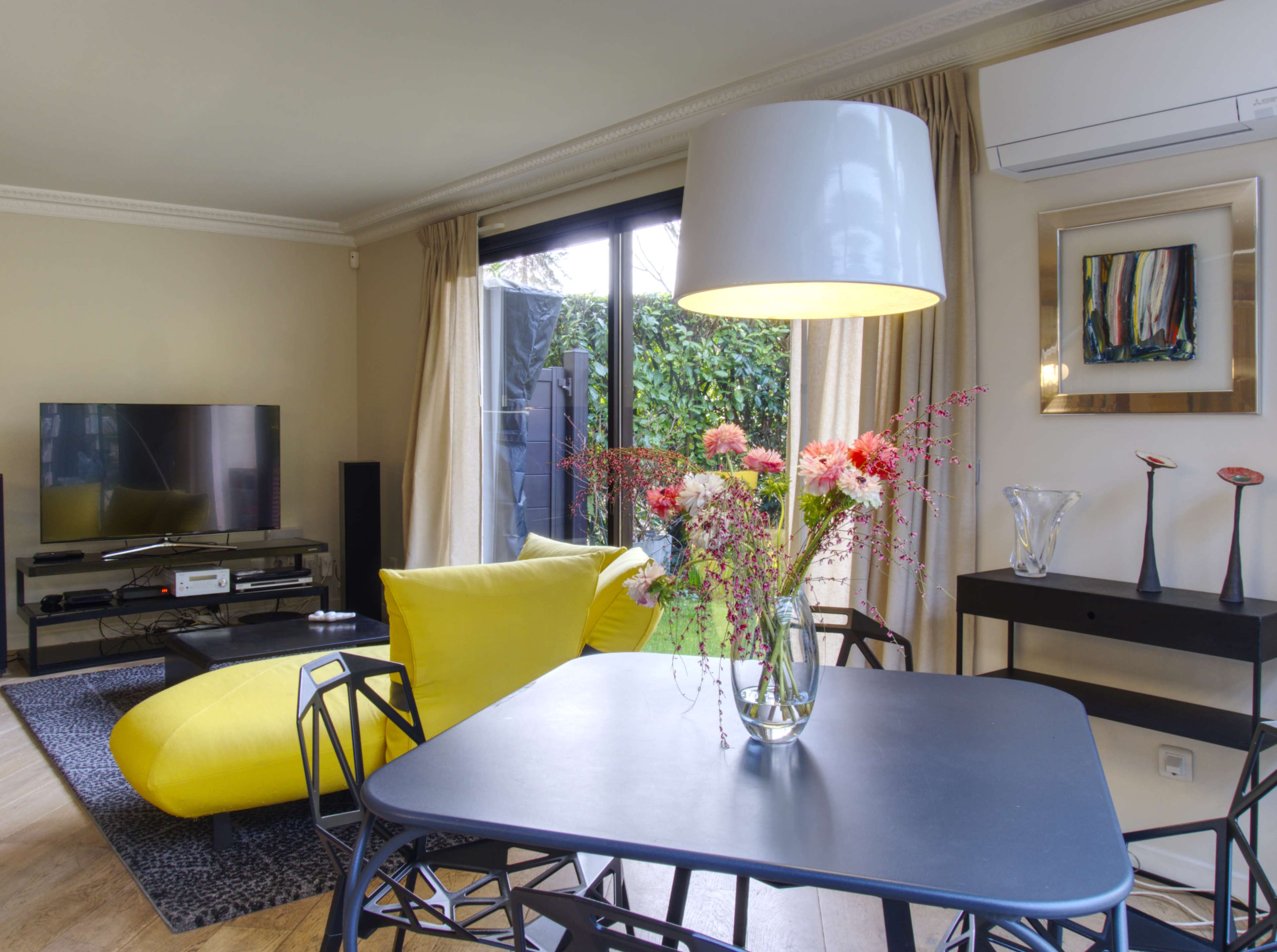 The image shows a modern living room with a yellow lounge chair, a dark dining table with flowers in a vase, a television, and large windows overlooking a garden.