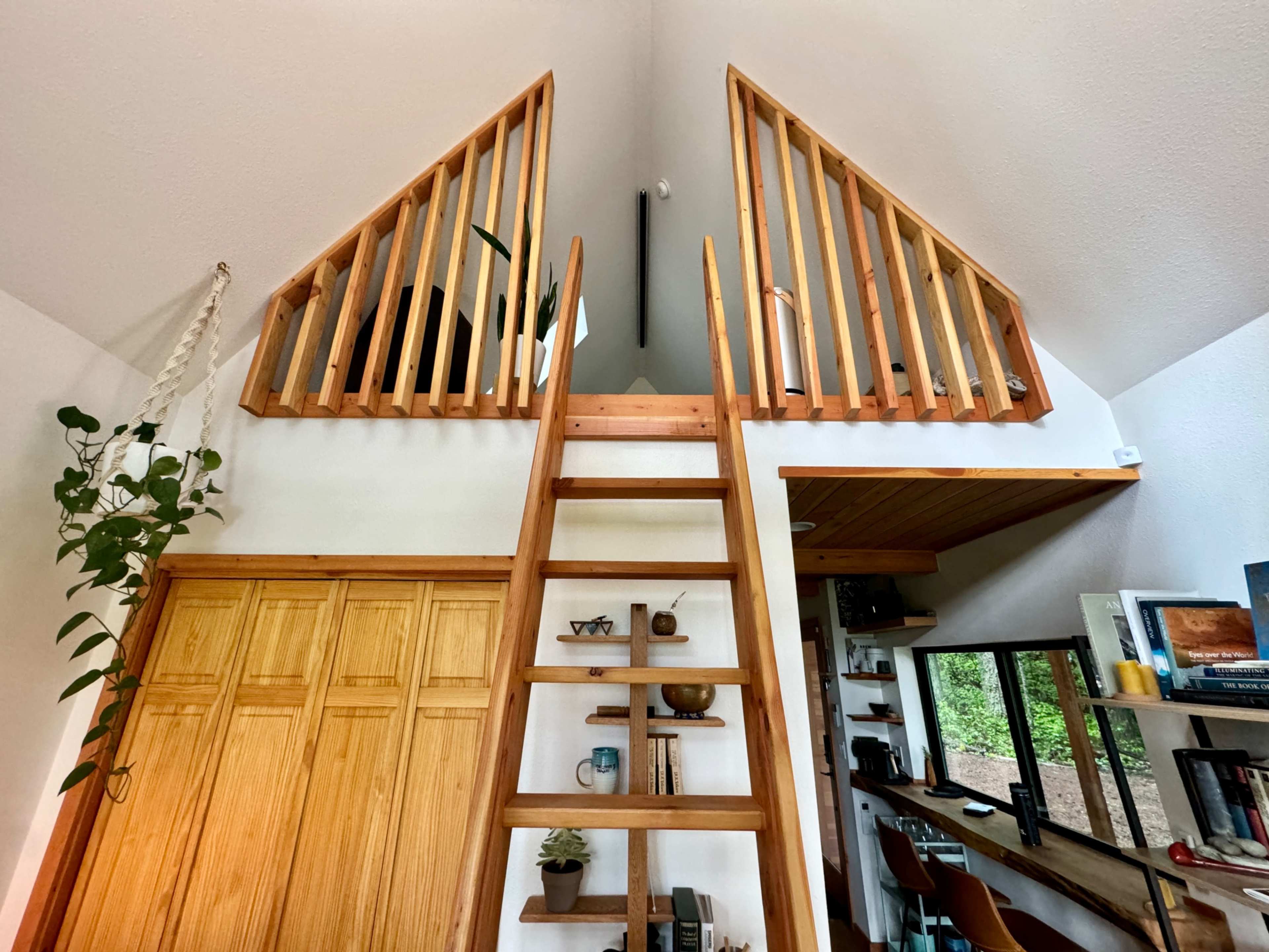 The image shows a wooden ladder leading to a loft area with railings, surrounded by white walls and wooden cabinetry in an open-concept space.