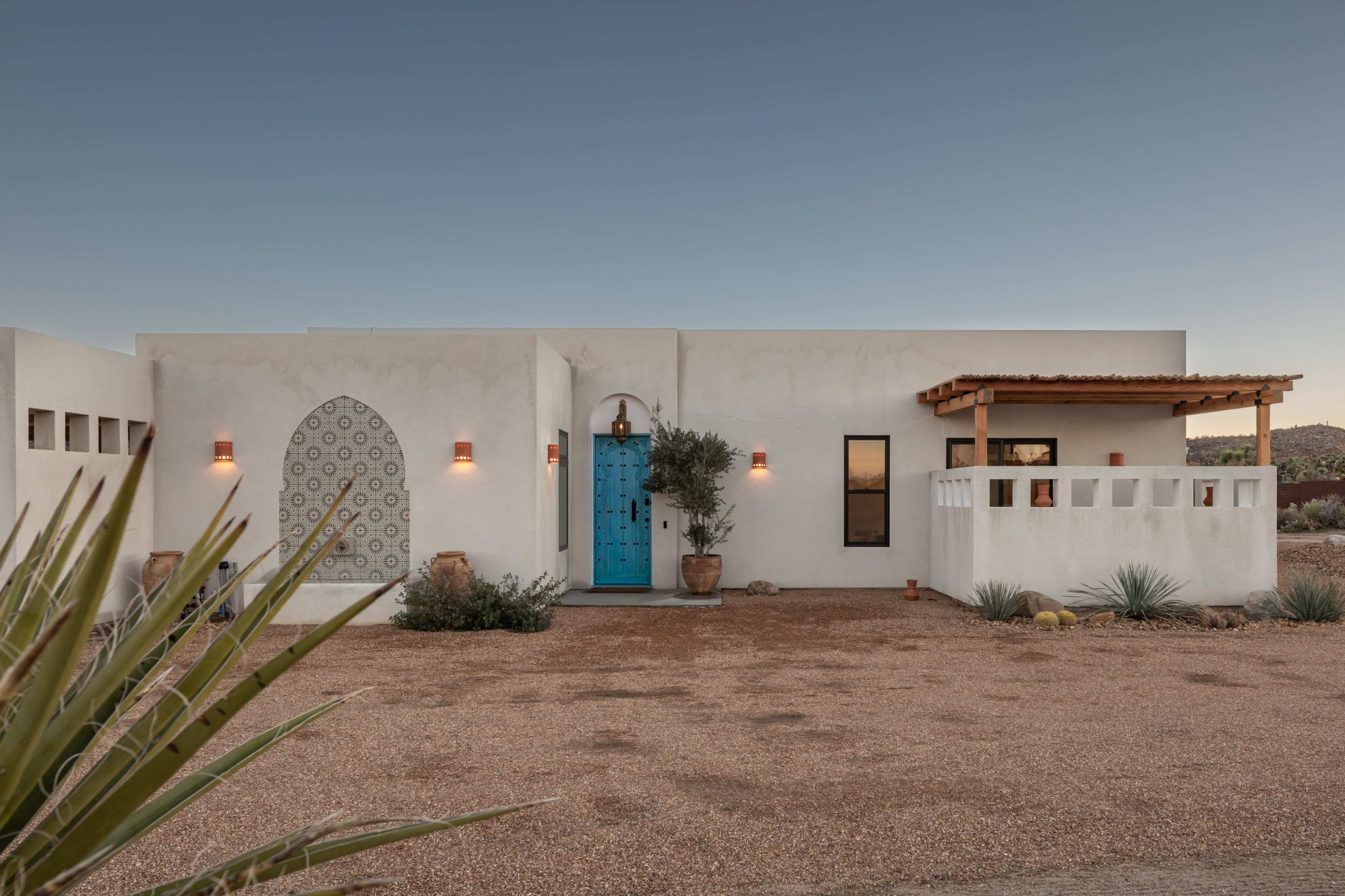 A modern adobe-style house with a turquoise door is set against a clear sky, surrounded by desert landscaping.