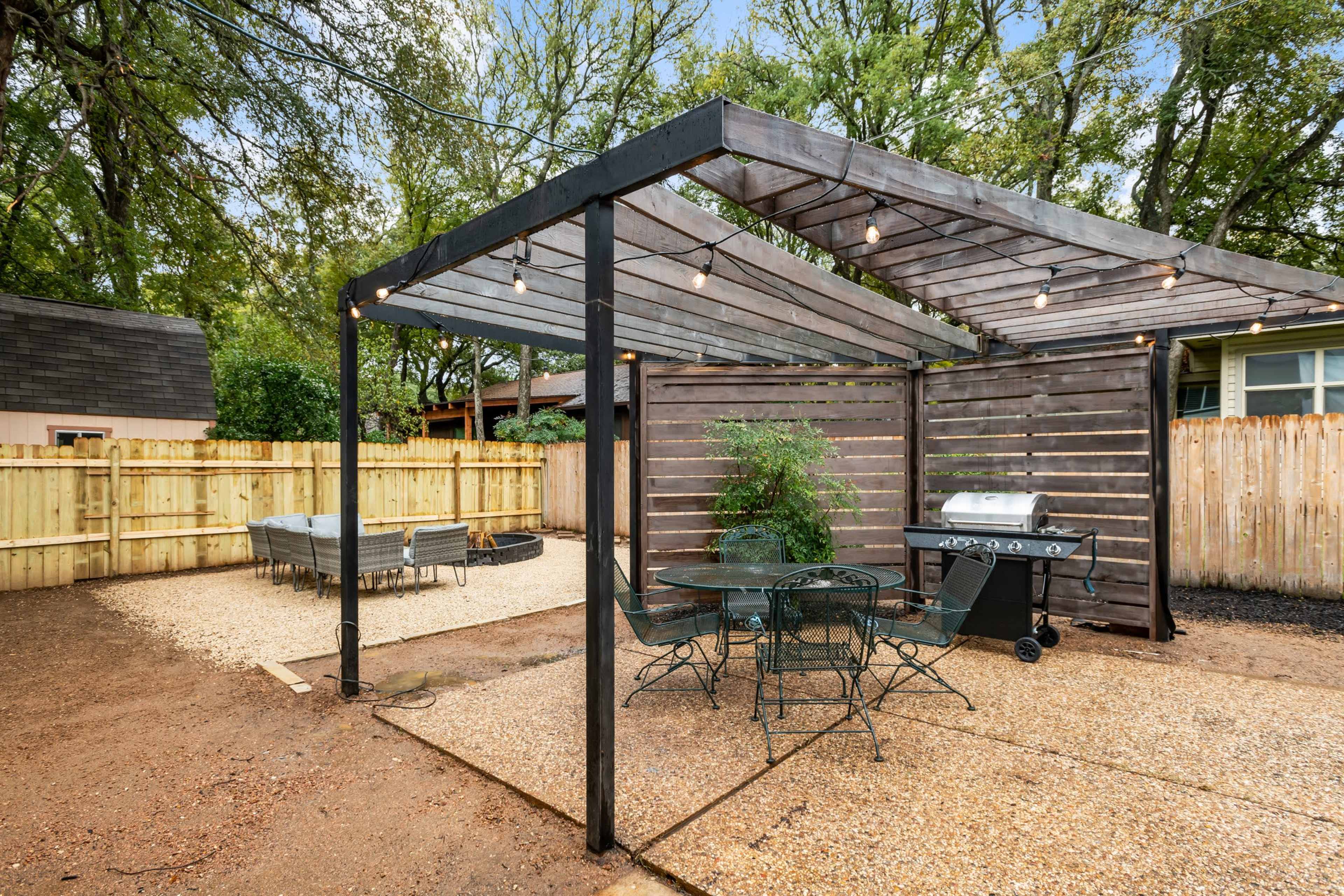 A structured outdoor patio area features a wooden pergola with string lights, a grill, and a dining set on a gravel surface surrounded by a wooden fence.