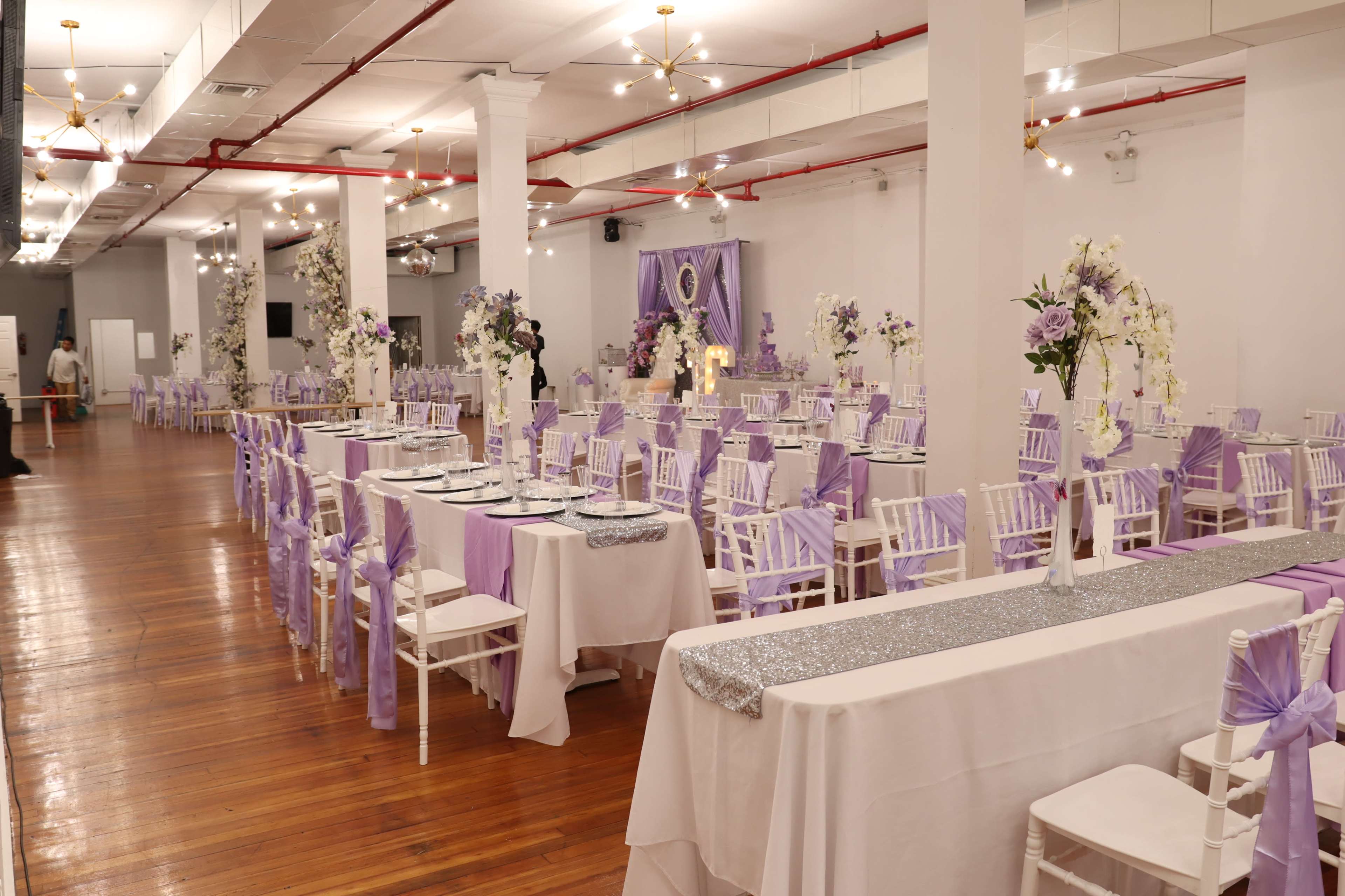 A large event hall is set up with white chairs adorned with lavender covers and elegantly decorated tables featuring lavender and silver accents.