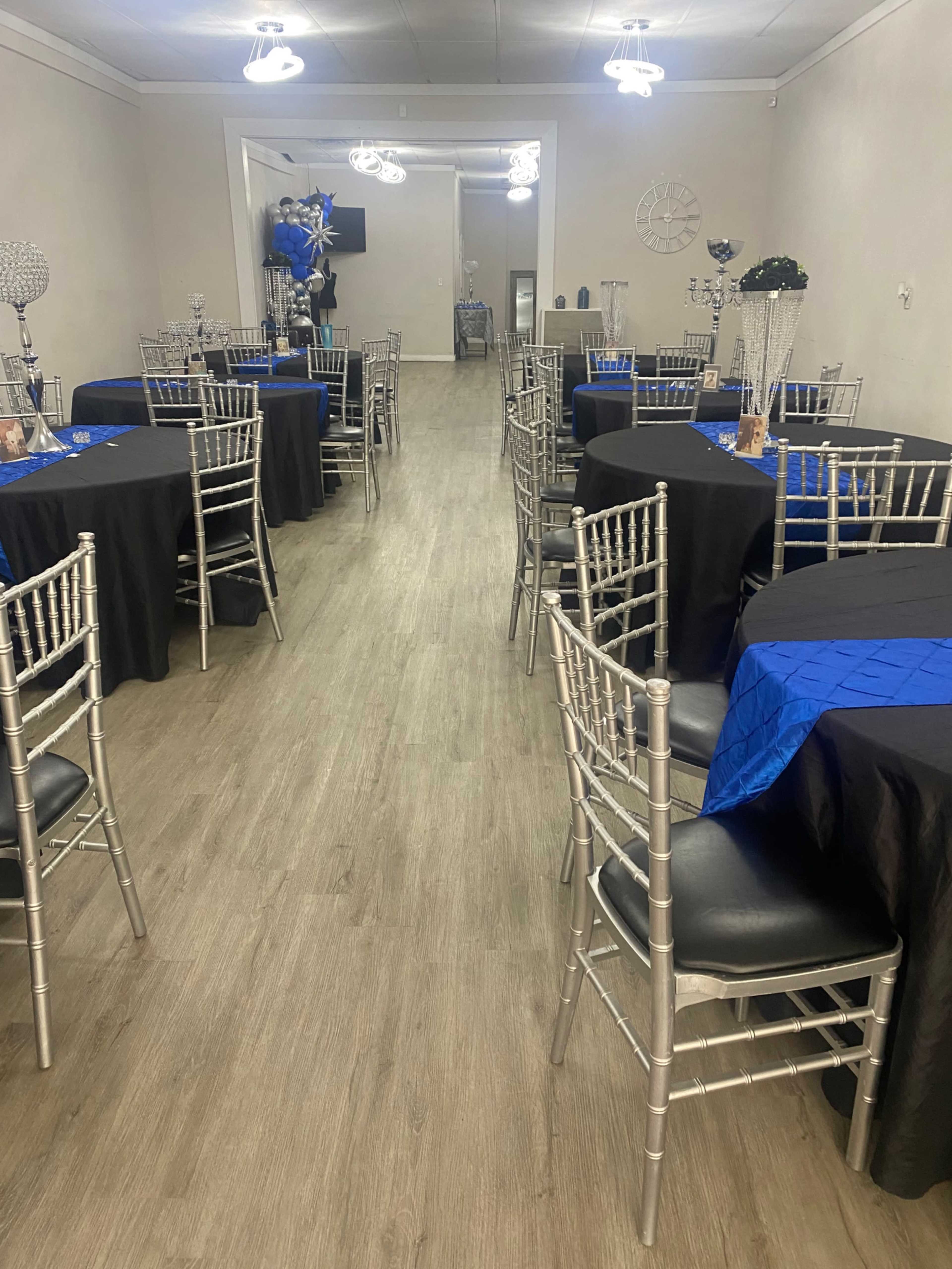 A spacious event room features tables covered with black linens and blue accents, surrounded by silver chiavari chairs.