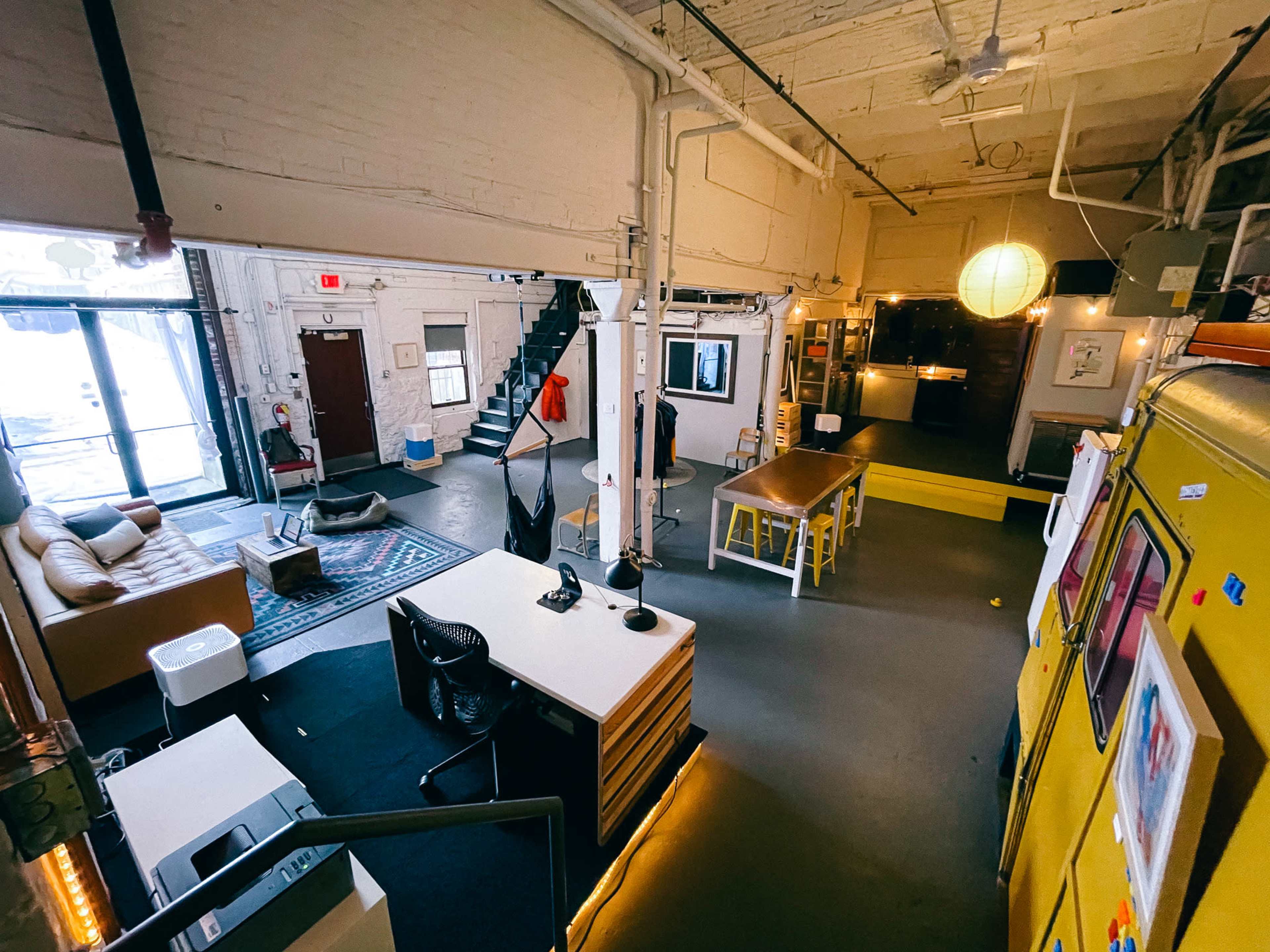 A spacious, industrial-style office with a seating area, a kitchen space, and a staircase leading to an upper level.