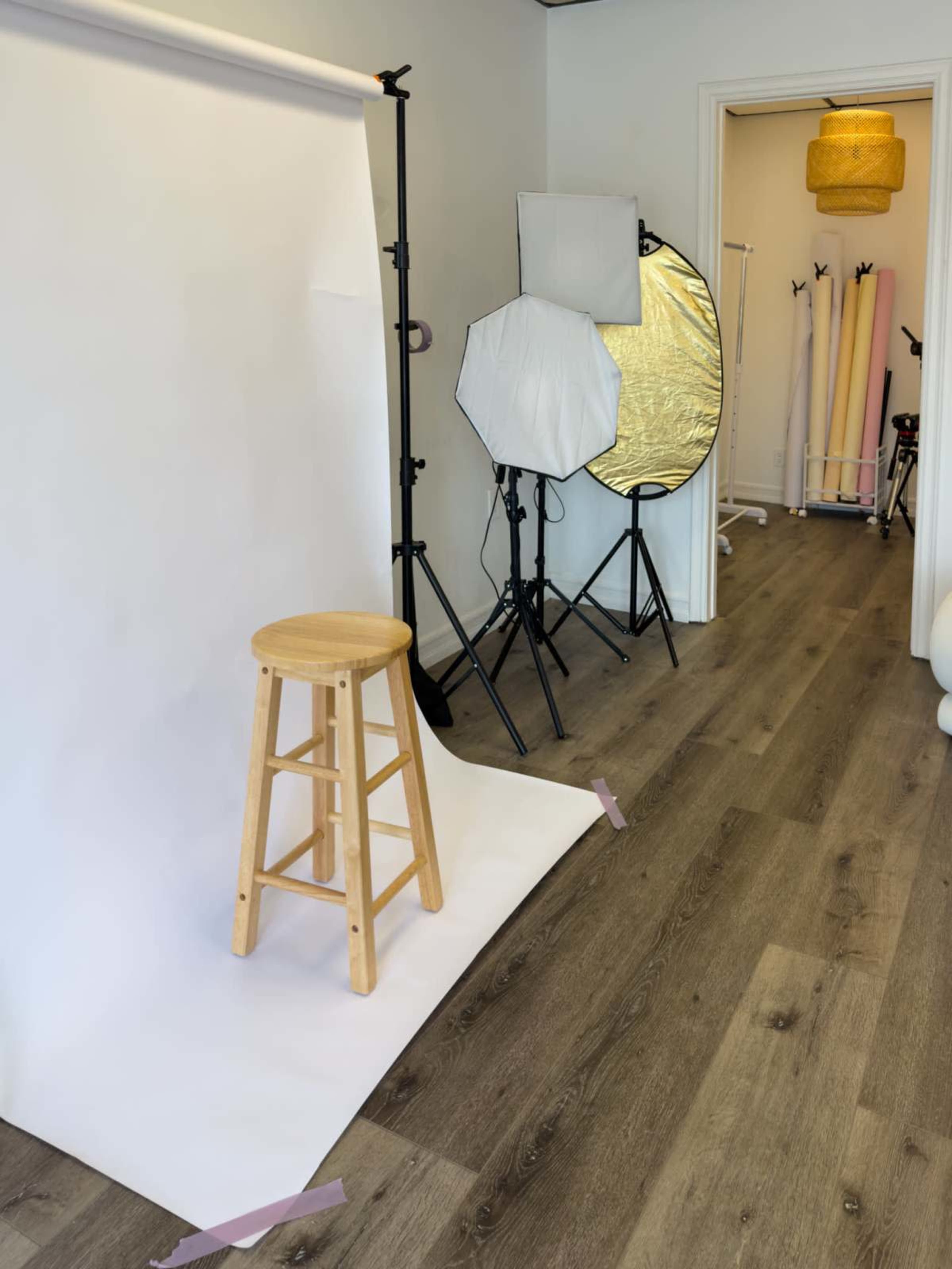 The image shows a photography studio setup featuring a wooden stool in front of a white backdrop, with various lighting equipment and reflectors arranged in the background.