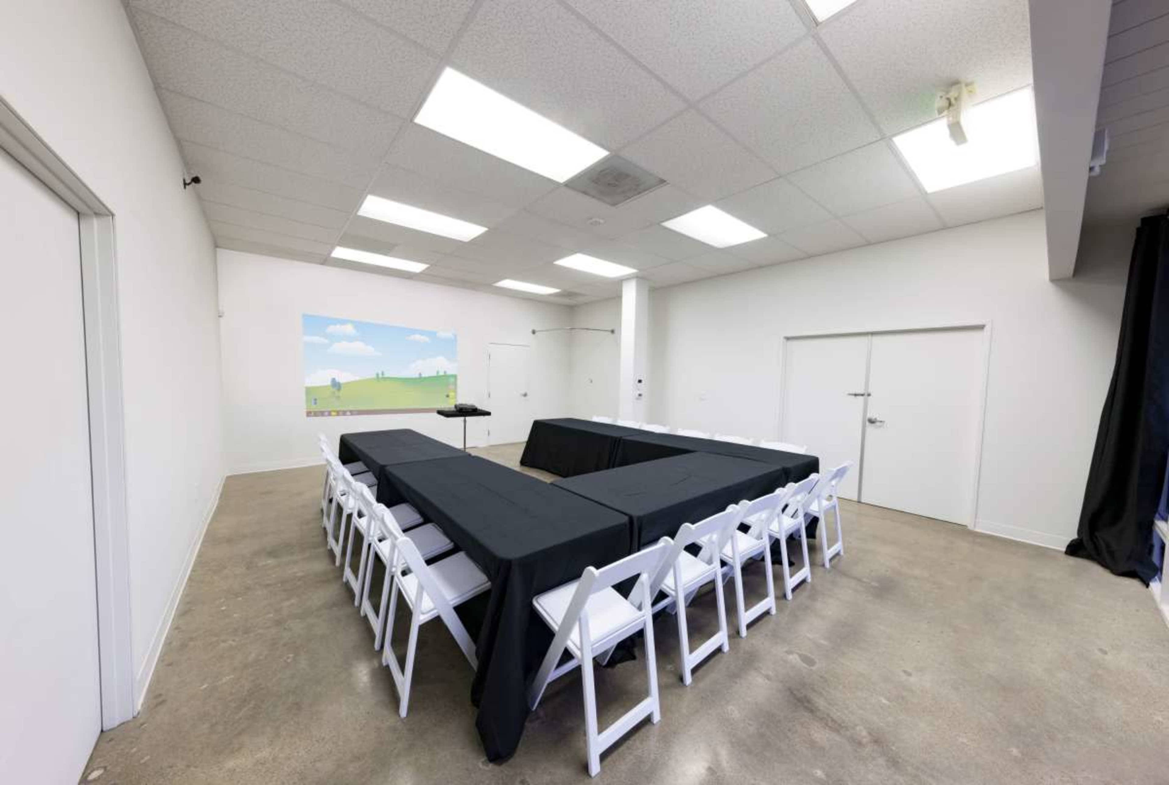 A rectangular arrangement of tables covered with black cloth is set up in a well-lit room with white walls and a projected image on one wall.