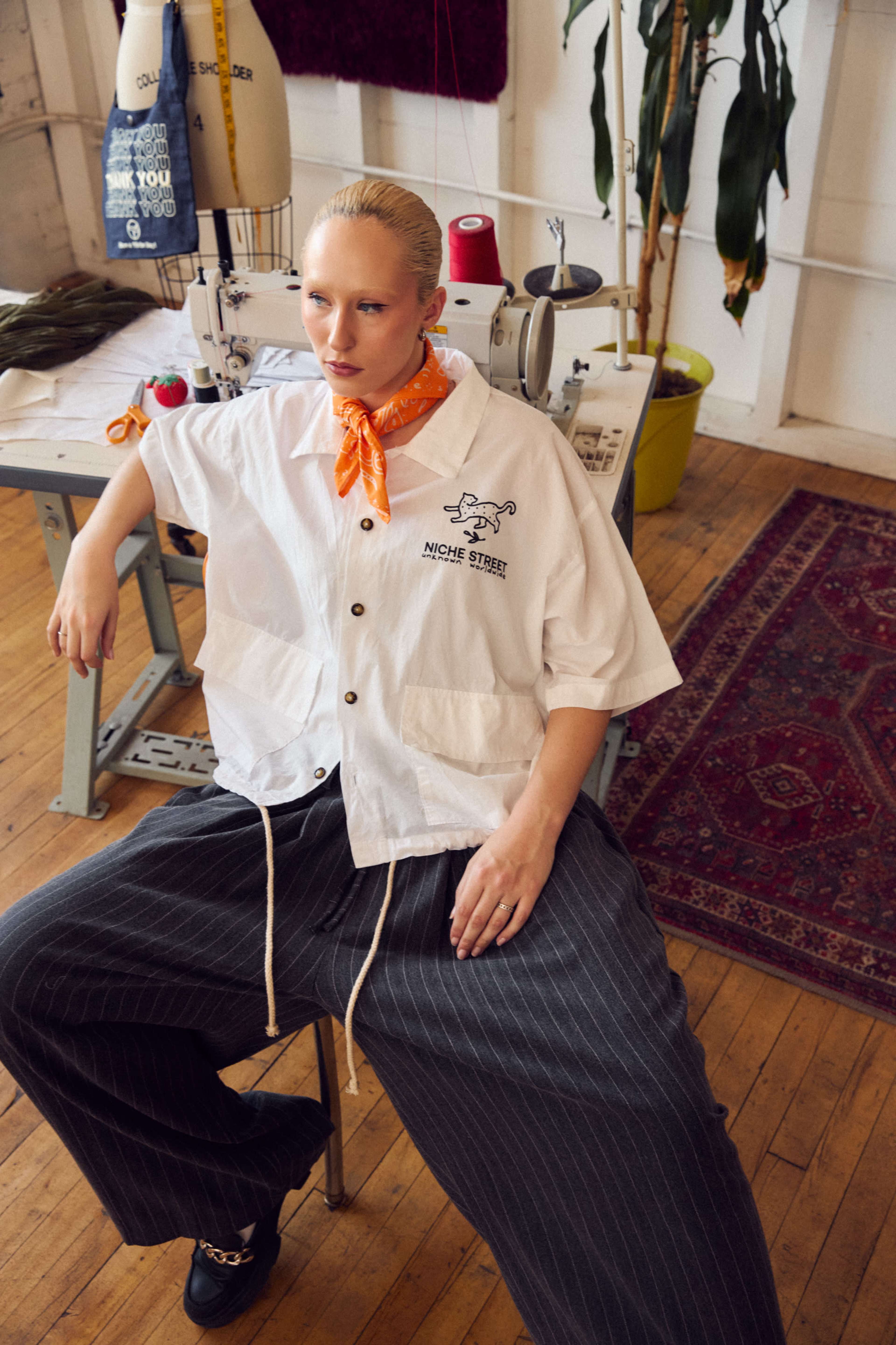 A person sits on a chair in a sewing studio, wearing a white button-up shirt with an orange neck scarf and loose-striped pants.