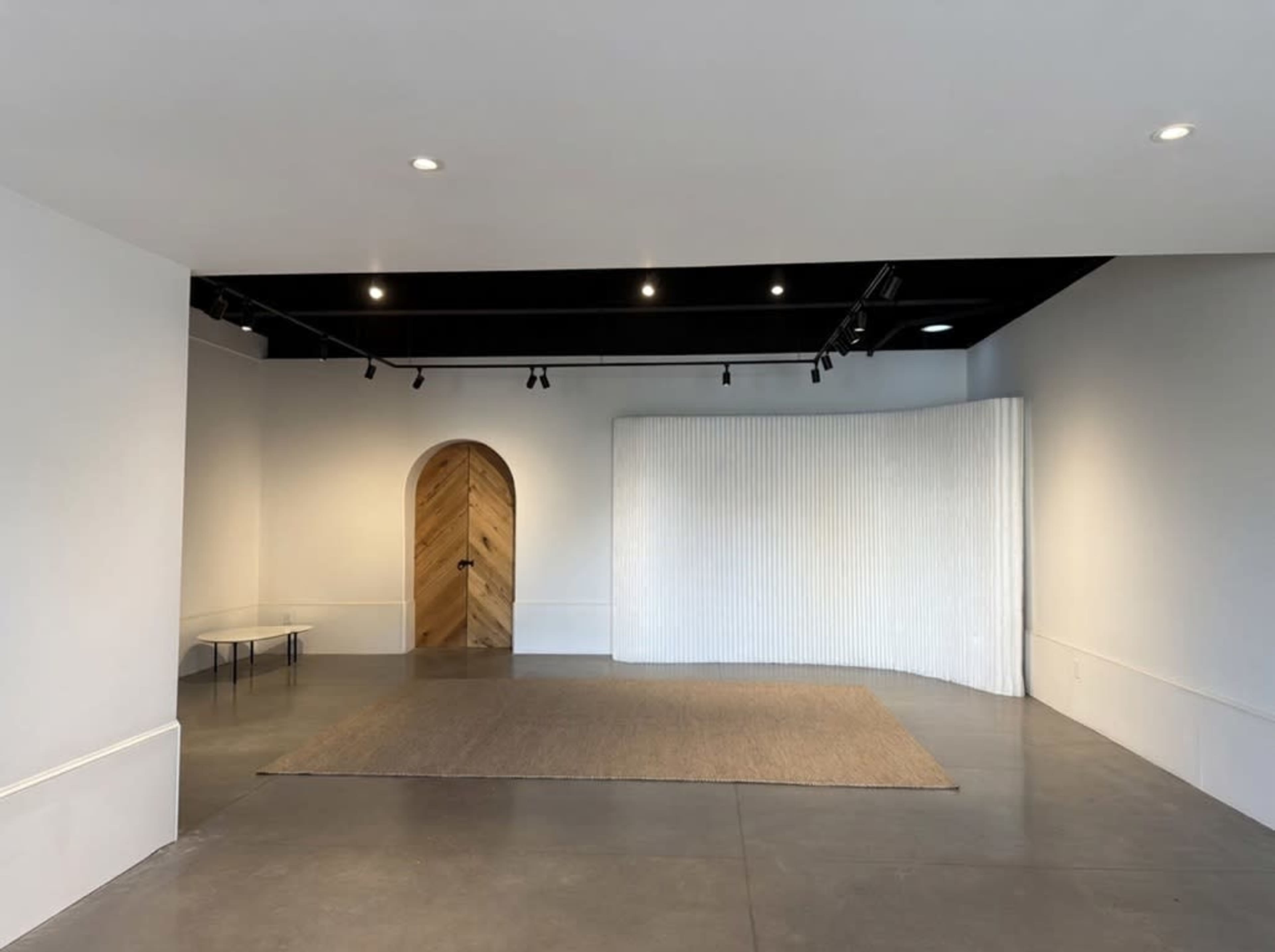 The image shows a minimalist room with a curved white wall, a wooden door with a herringbone pattern, a rectangular rug, and a table in the corner.
