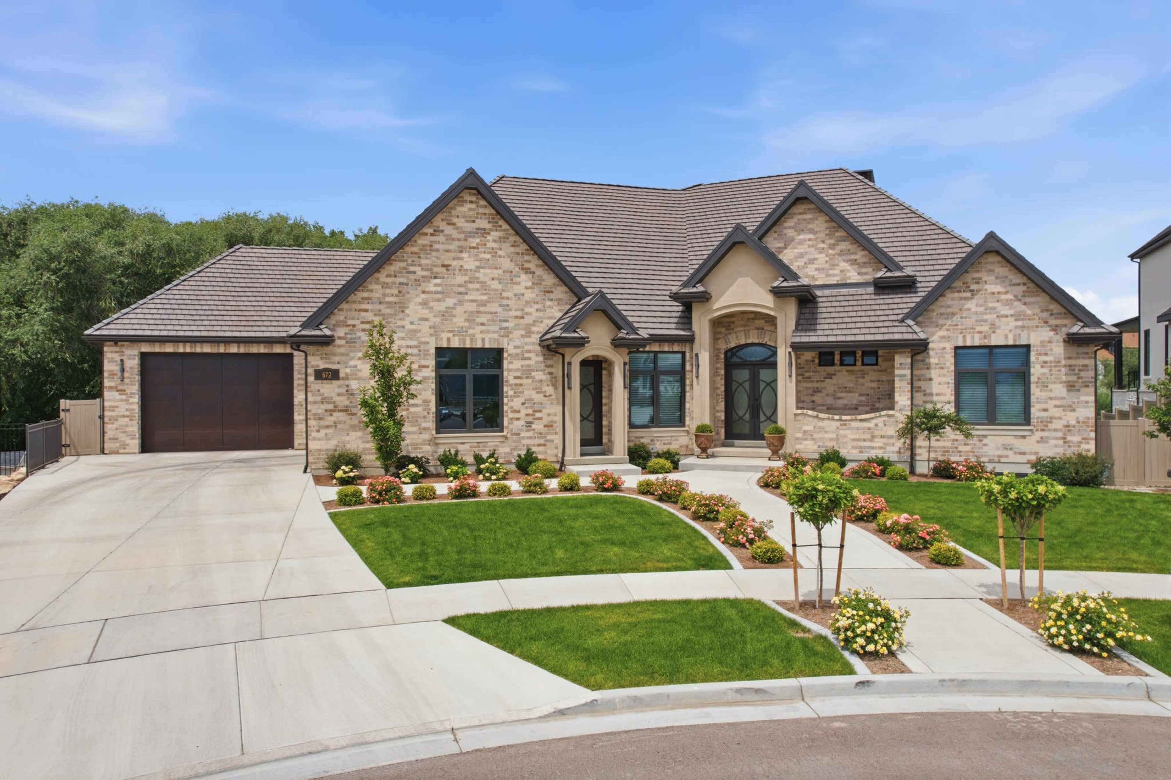 A single-story brick house with a gabled roof is situated on a landscaped corner lot with a curved driveway and neatly arranged flower beds.