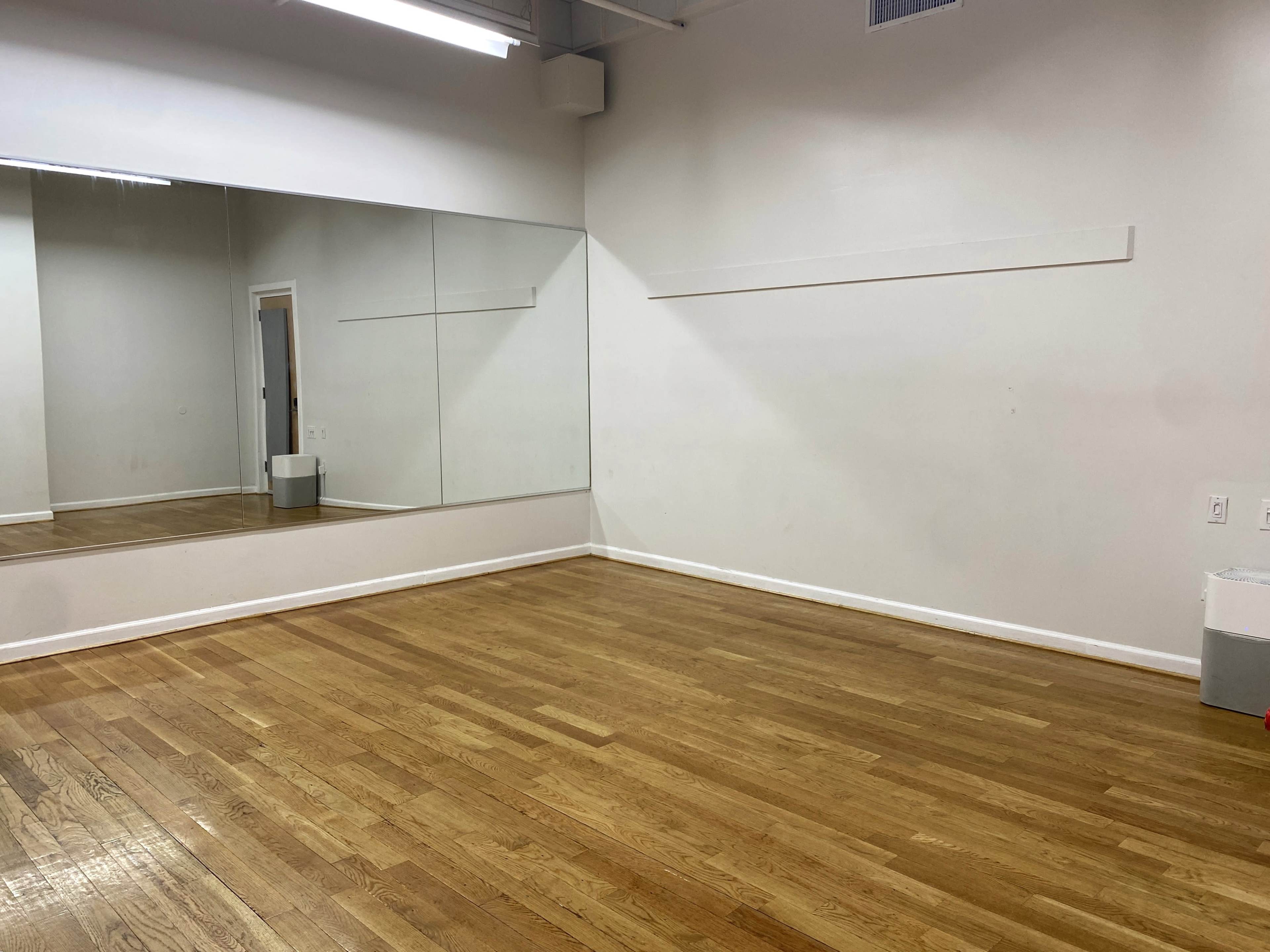 The image shows a spacious room with wooden floors, a wall mirror, and minimal furnishings.