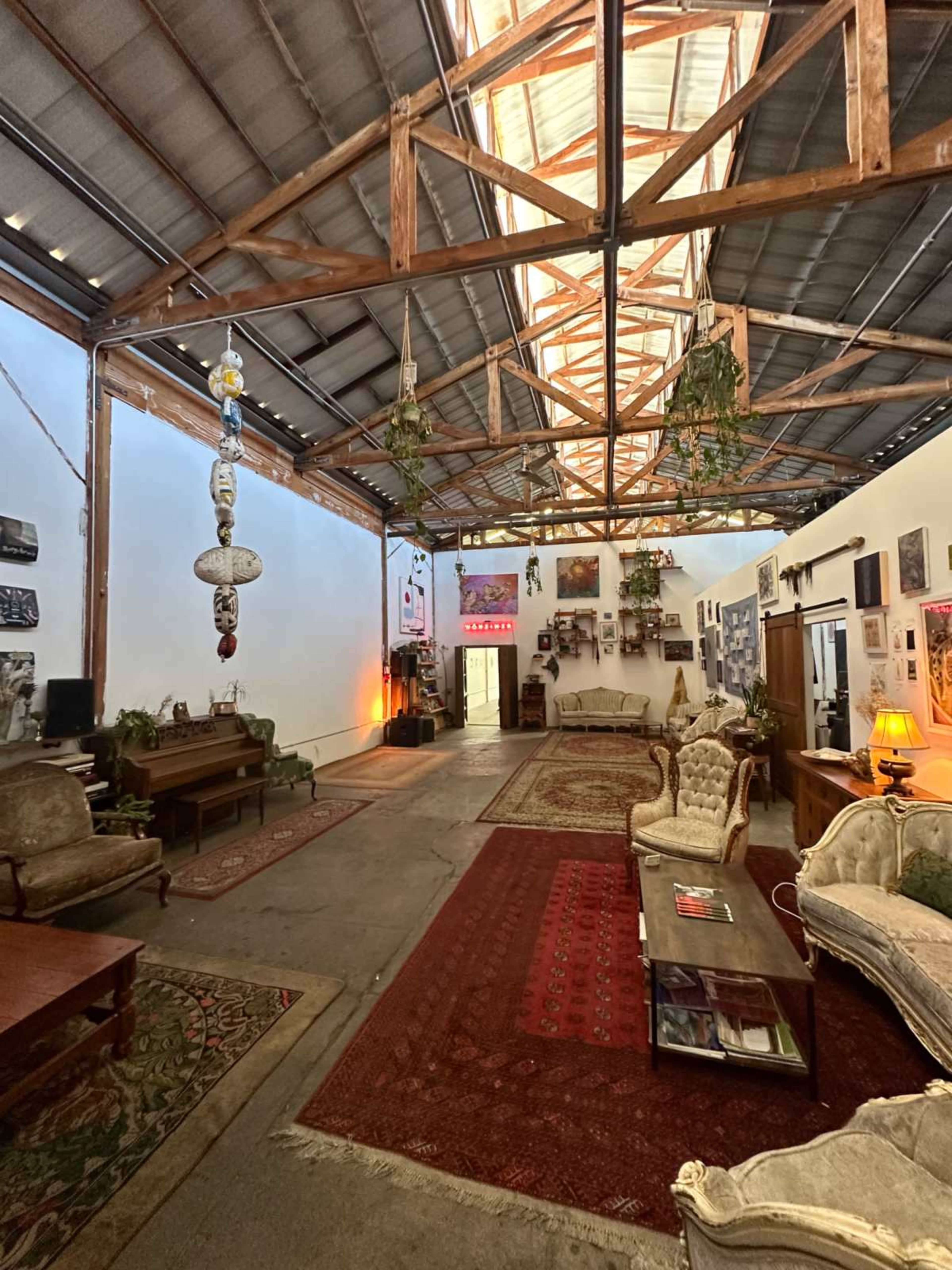 The image shows a spacious, industrial-style room with wooden beams, vintage furniture, rugs on the floor, and various decorations on the walls.