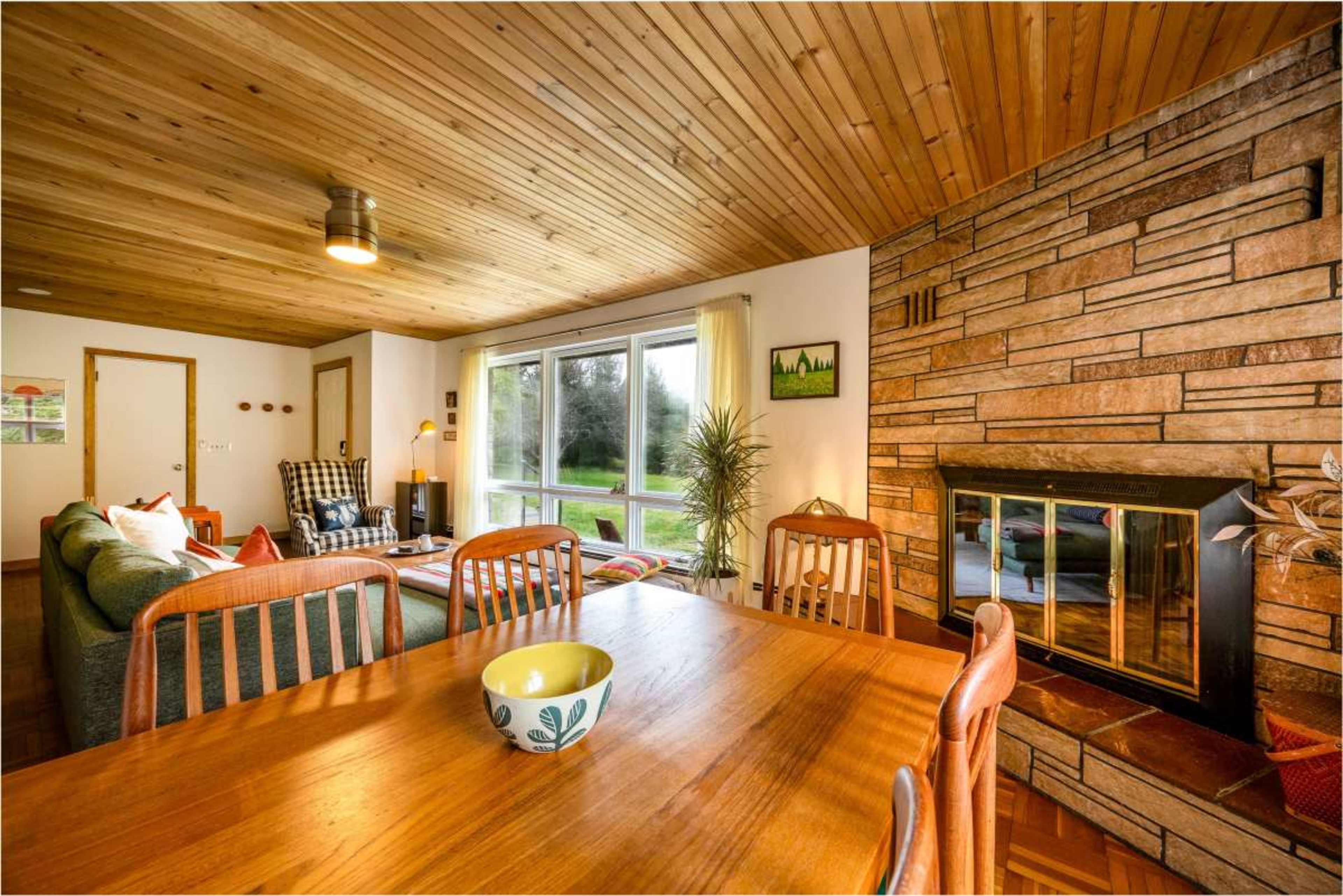 A wooden-paneled living area features a dining table, a sofa set, a fireplace, and large windows overlooking a green landscape.