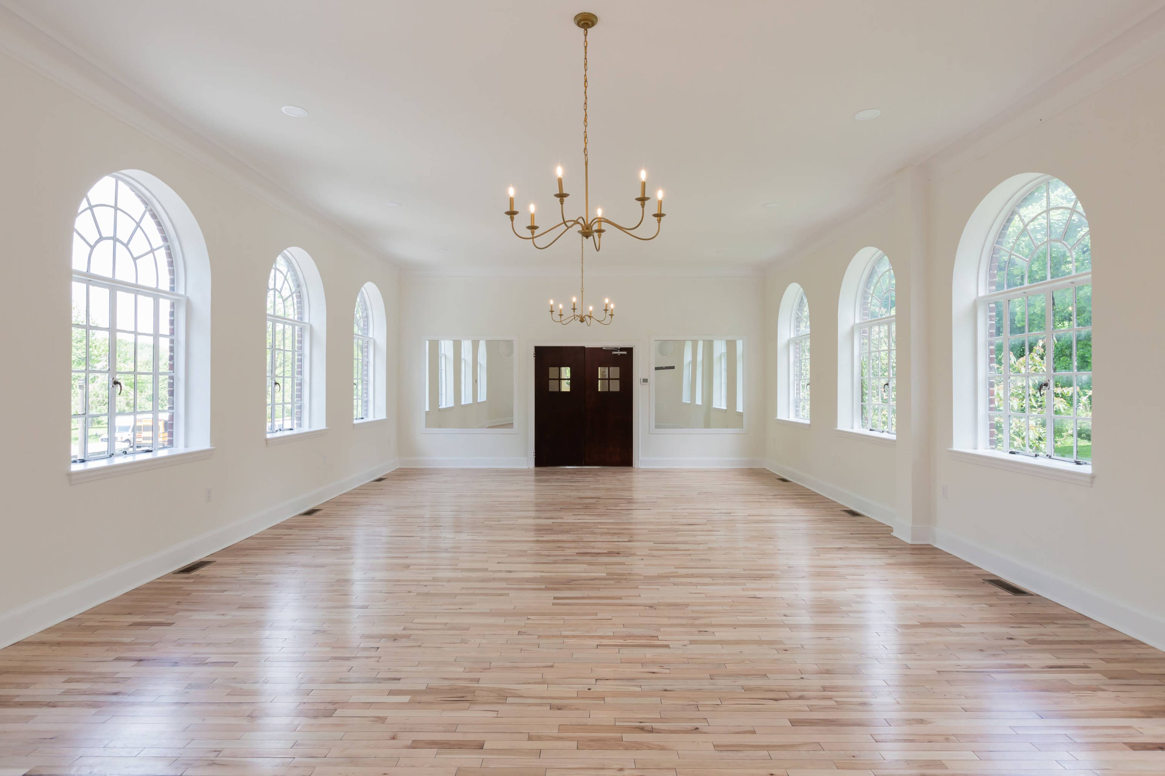 A spacious, well-lit room with hardwood floors, large arched windows, and a chandelier overhead, leading to a set of double doors.
