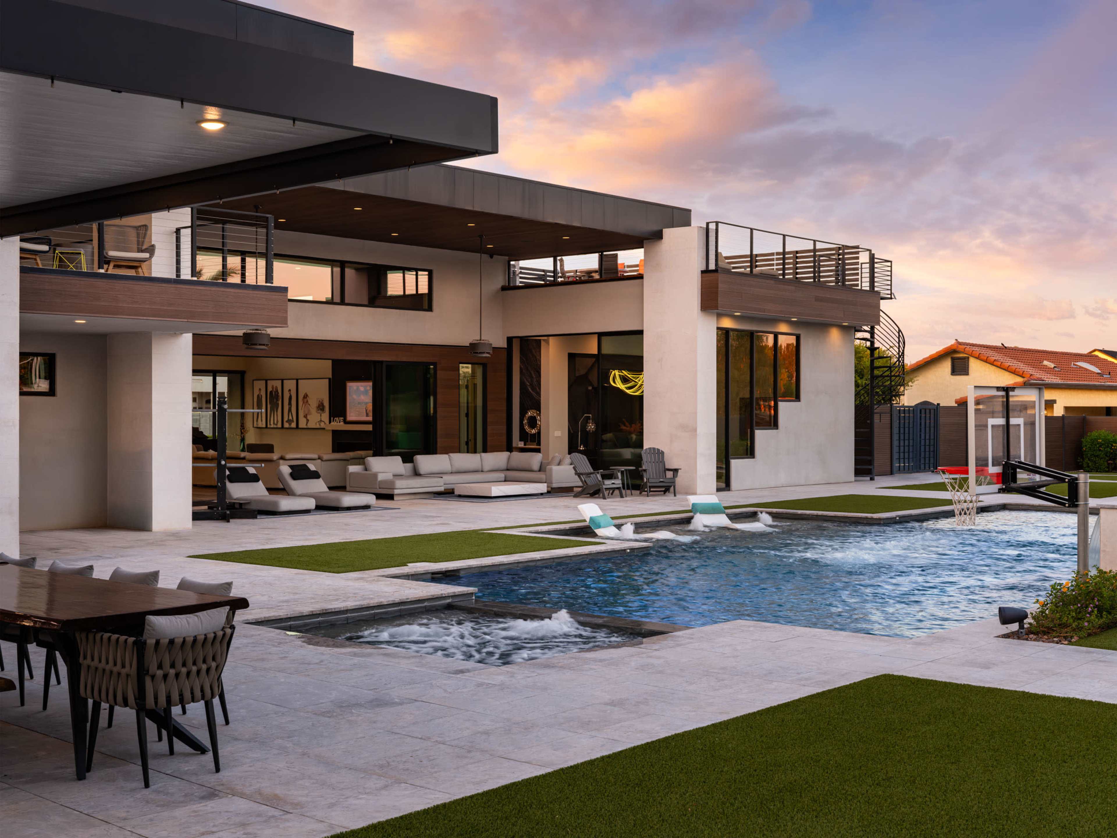 The image features a modern house with a spacious outdoor area that includes a swimming pool, lounge seating, and a basketball hoop amidst manicured grass.