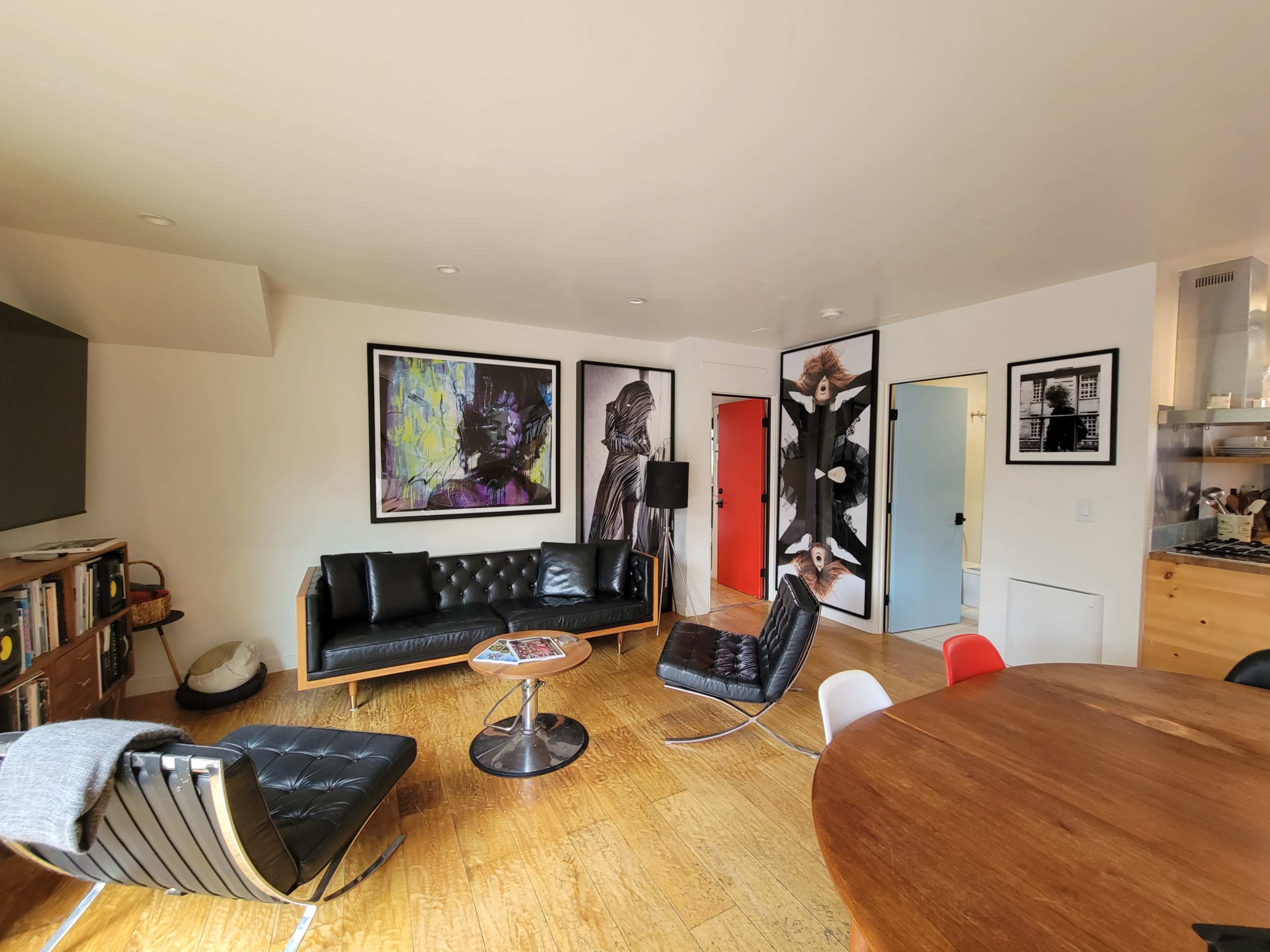 The image shows a modern living room with a black leather sofa, a round coffee table, and various artworks on the walls, alongside a dining table with colorful chairs.