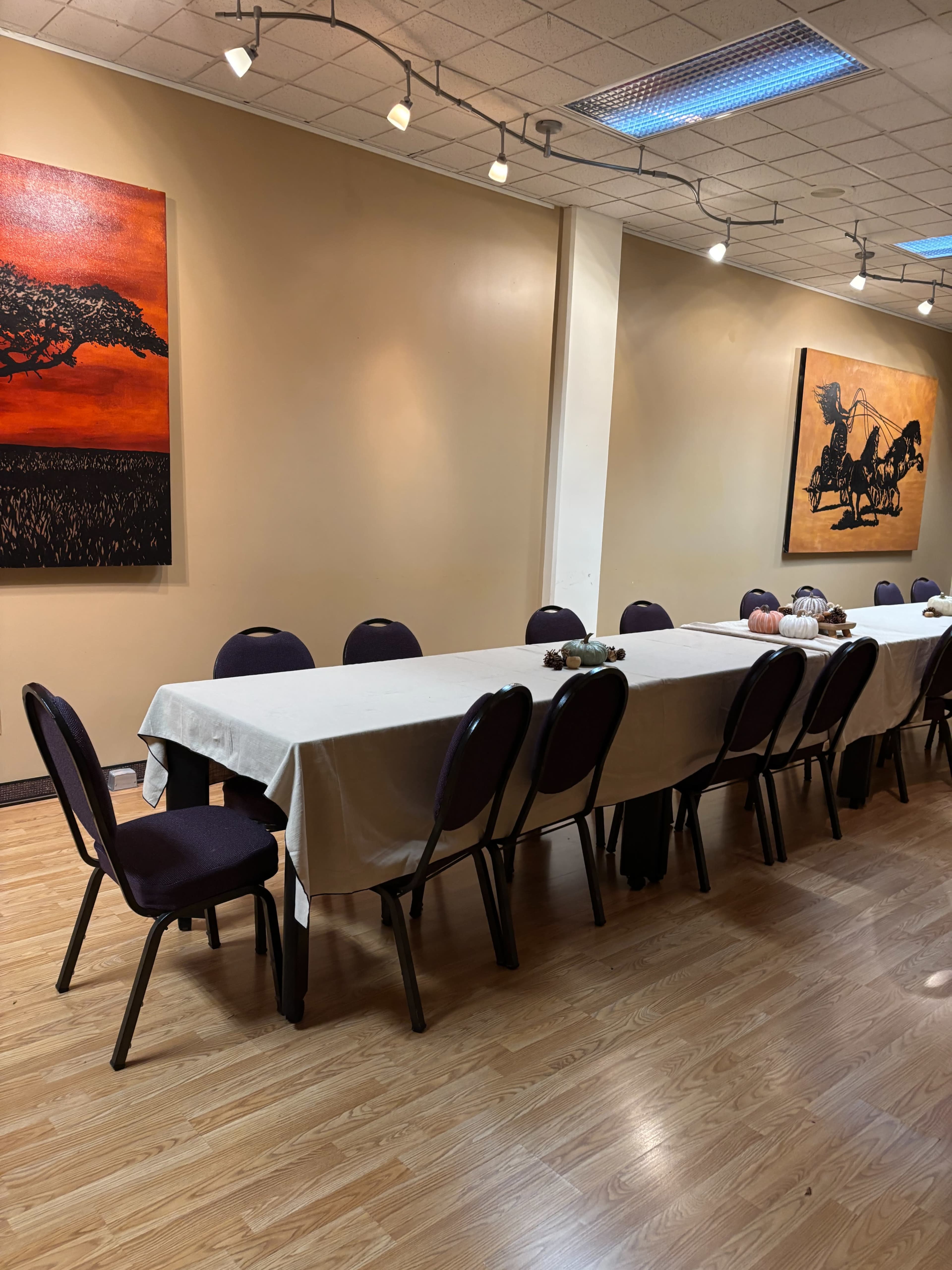 A long table covered with a white tablecloth is set in a sparsely decorated room featuring two large paintings on the walls.