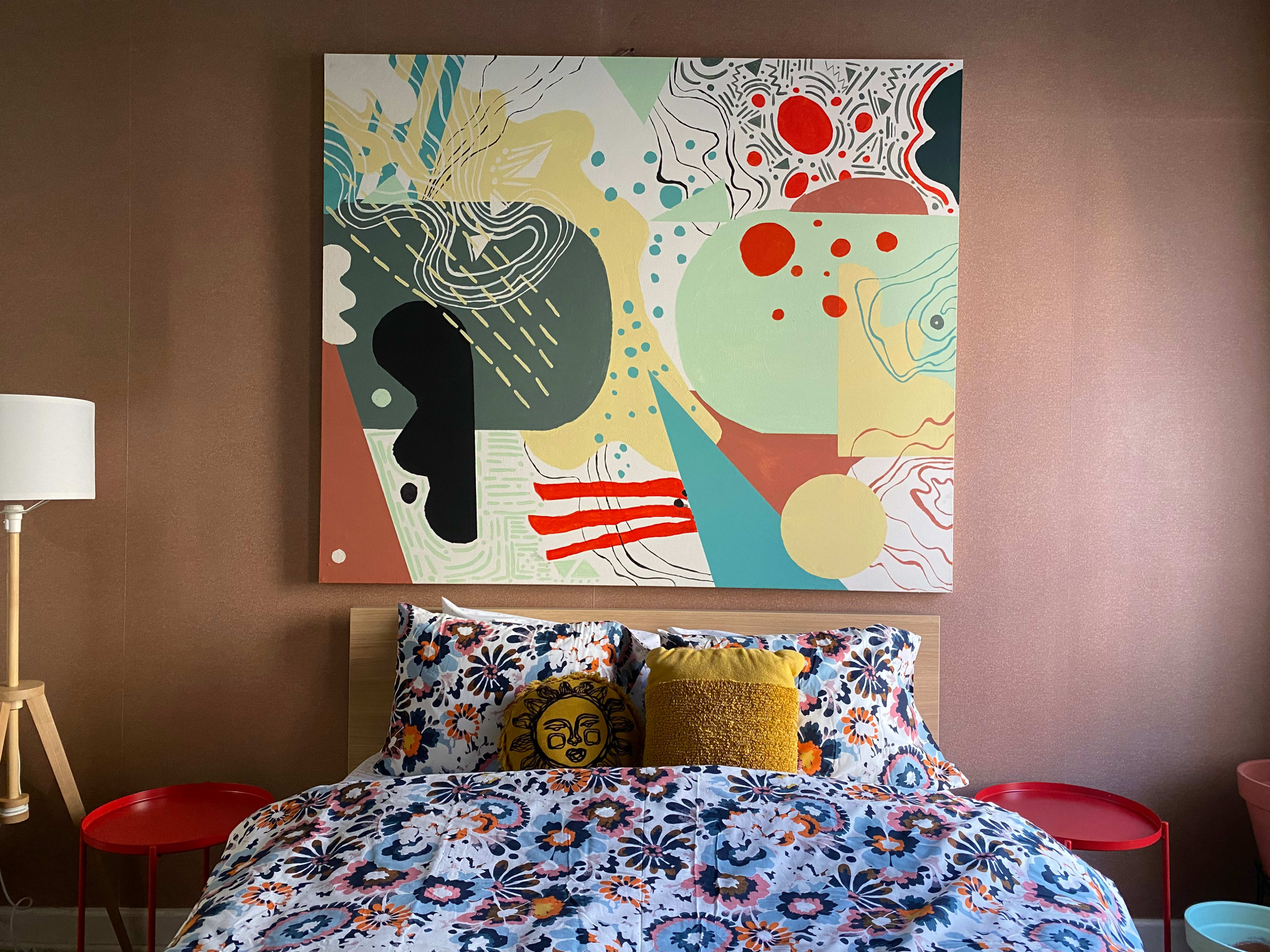 The image shows a bedroom featuring a large abstract painting above a wooden bed adorned with colorful bedding and decorative pillows, alongside two red bedside tables and a floor lamp.