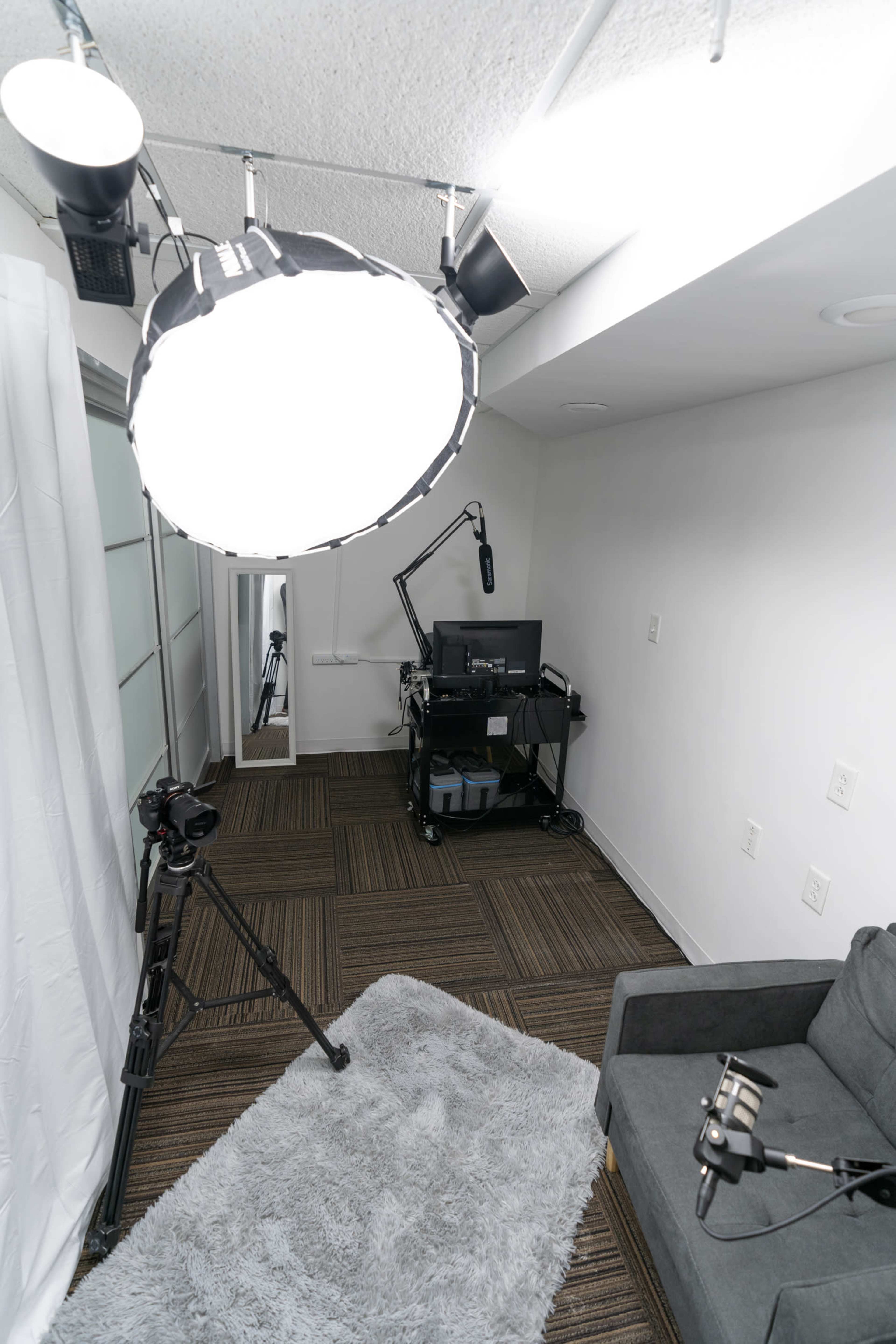 A small studio setup with a light fixture, camera equipment, a desk with a monitor, and a gray sofa on a rug.