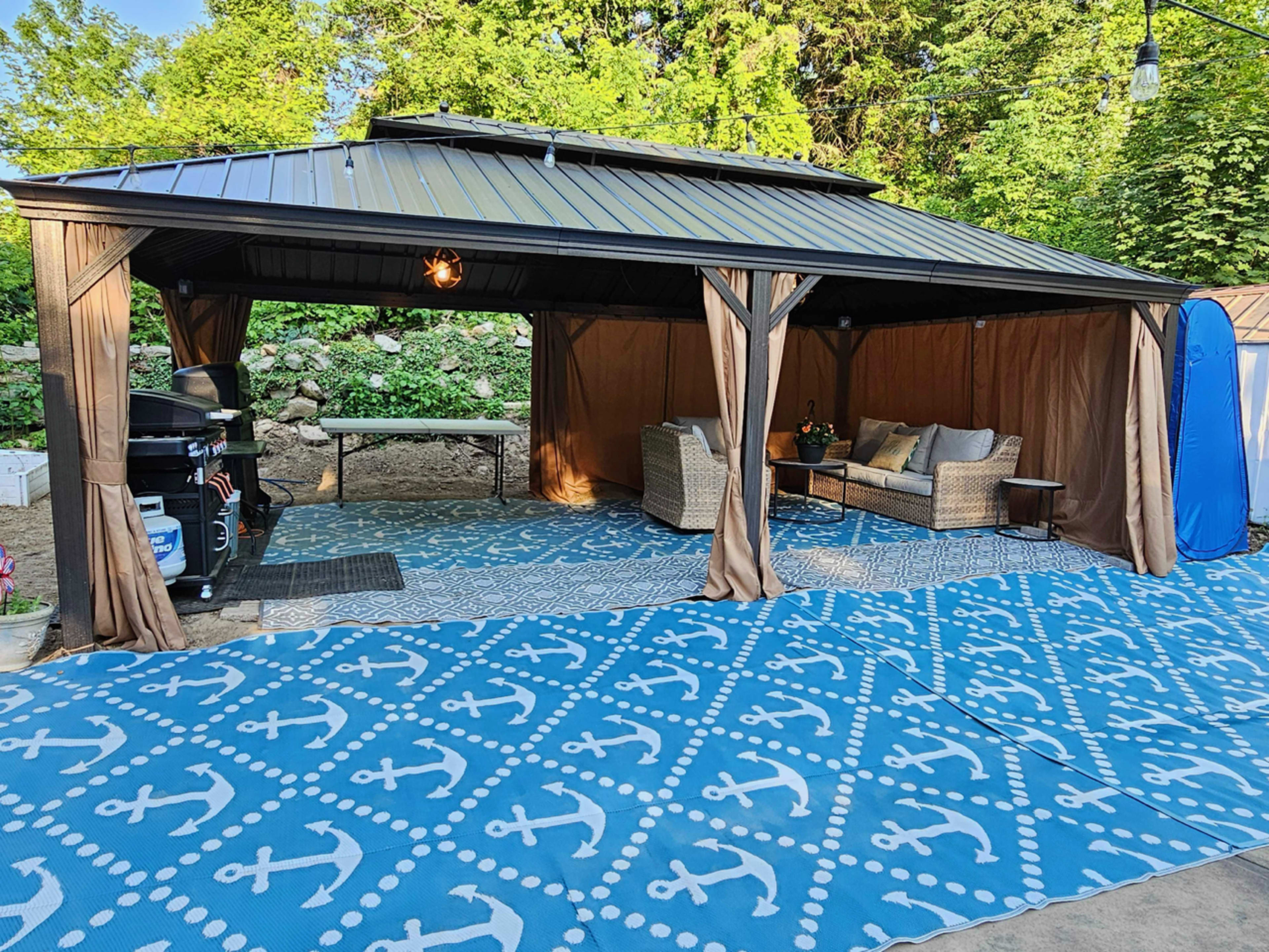 A covered outdoor seating area features beige furniture and a blue anchor-patterned rug, surrounded by greenery.