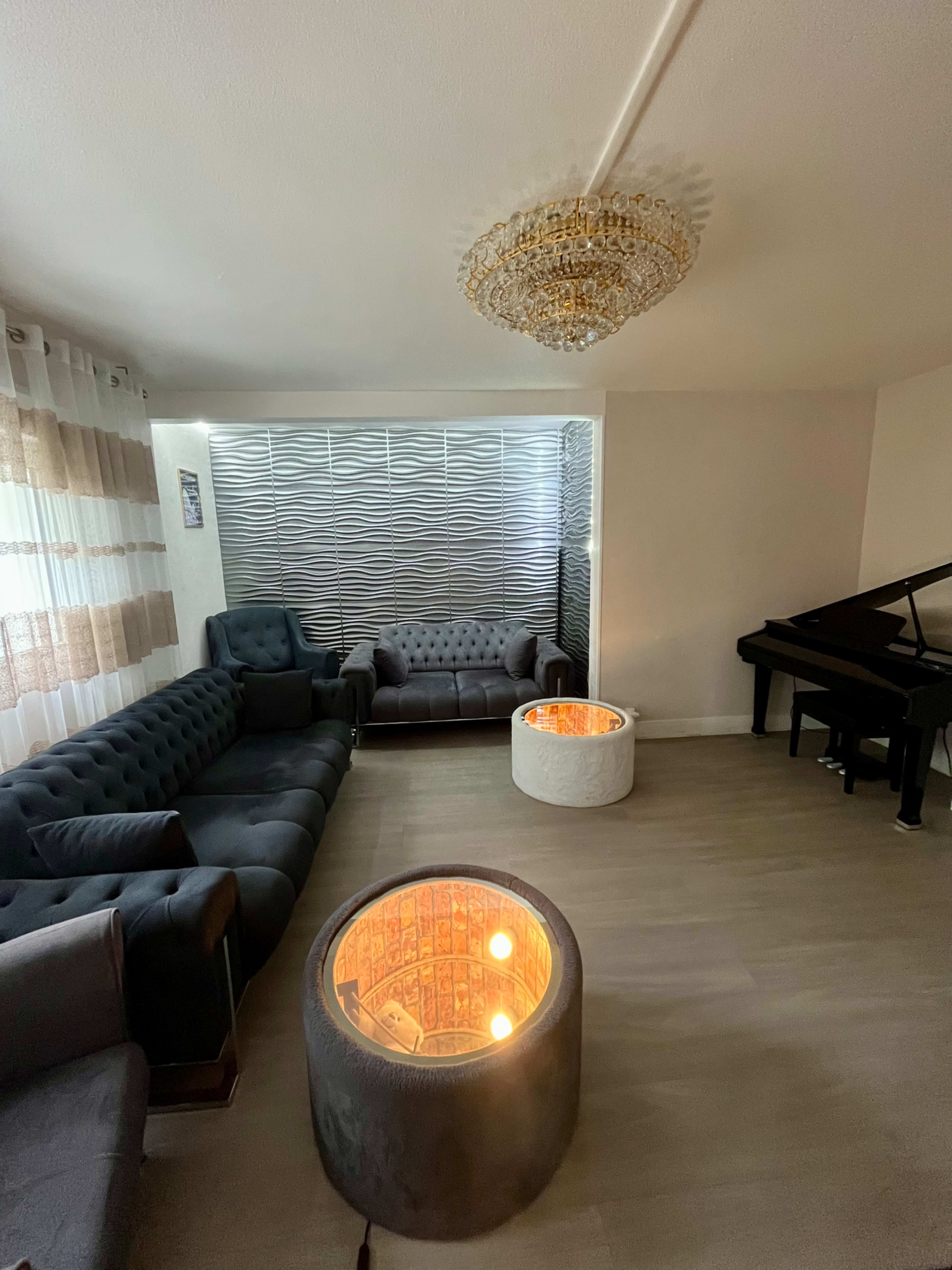 The image shows a modern living room featuring a dark sofa set, two illuminated circular tables, and a piano against a textured wall.