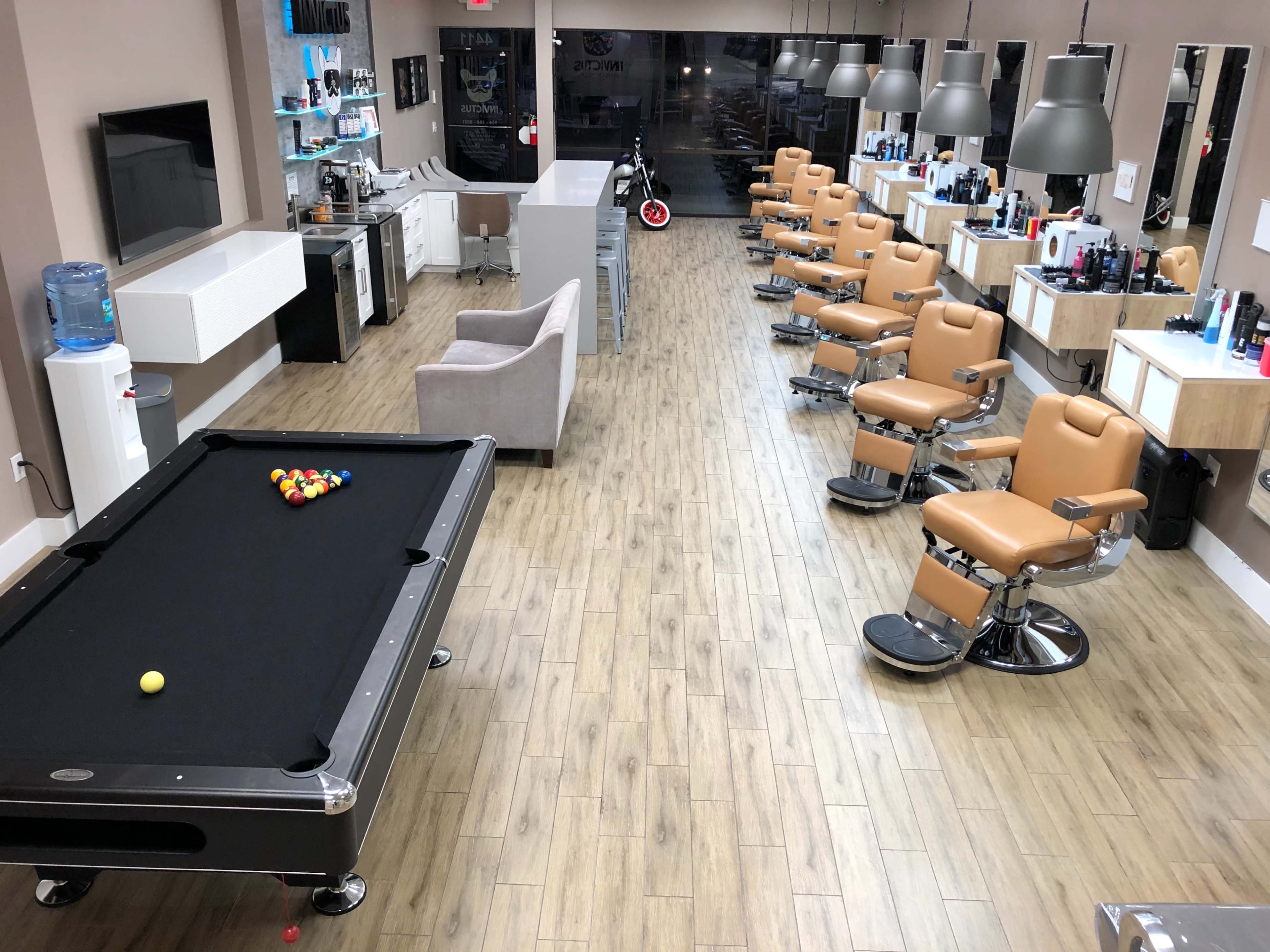 A spacious barbershop with several styling chairs lined up, a pool table in the foreground, and various grooming products displayed on counters.