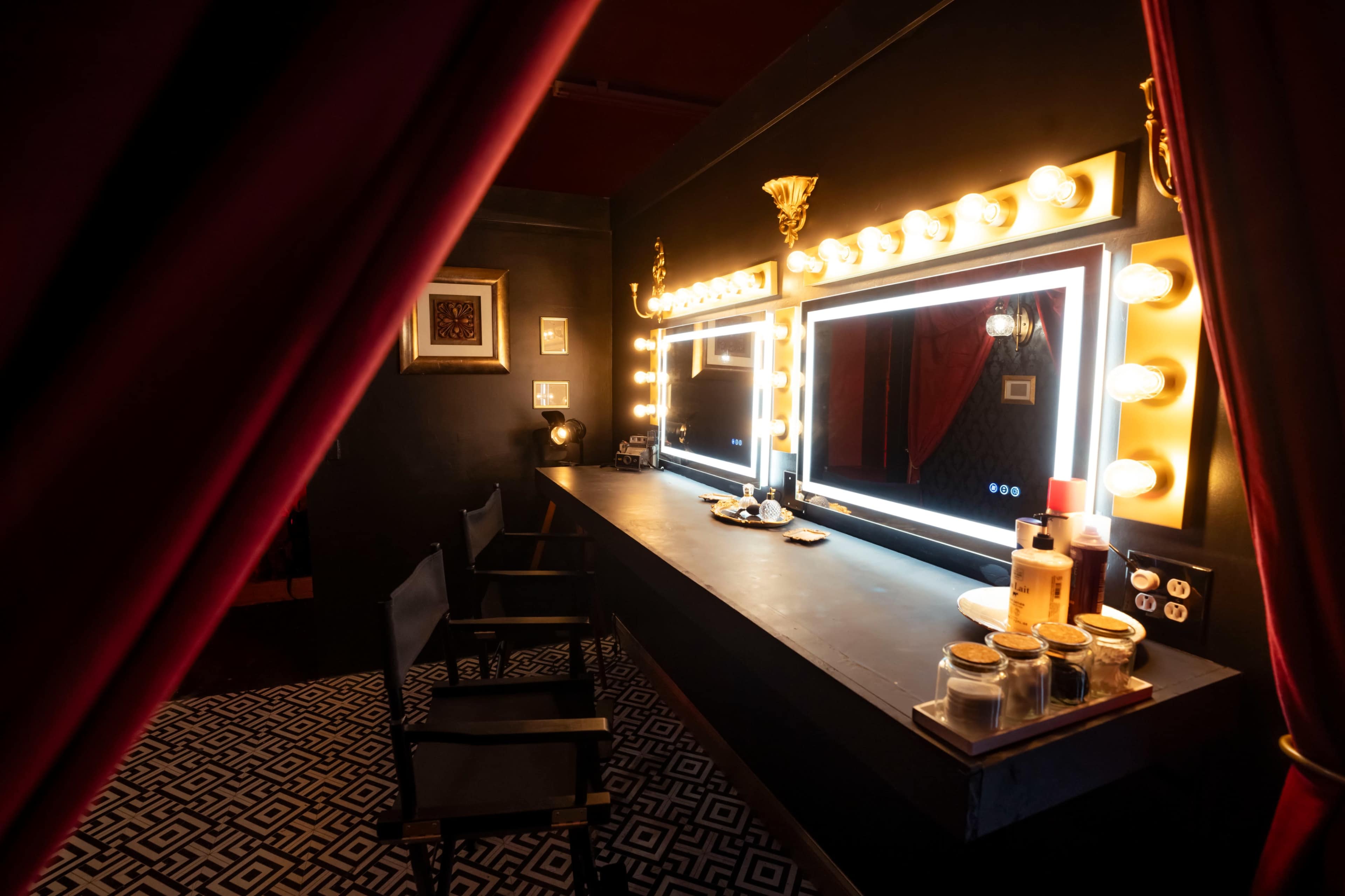 The image shows a dimly lit dressing room with a long black table, two illuminated mirrors, and a backdrop of red curtains.
