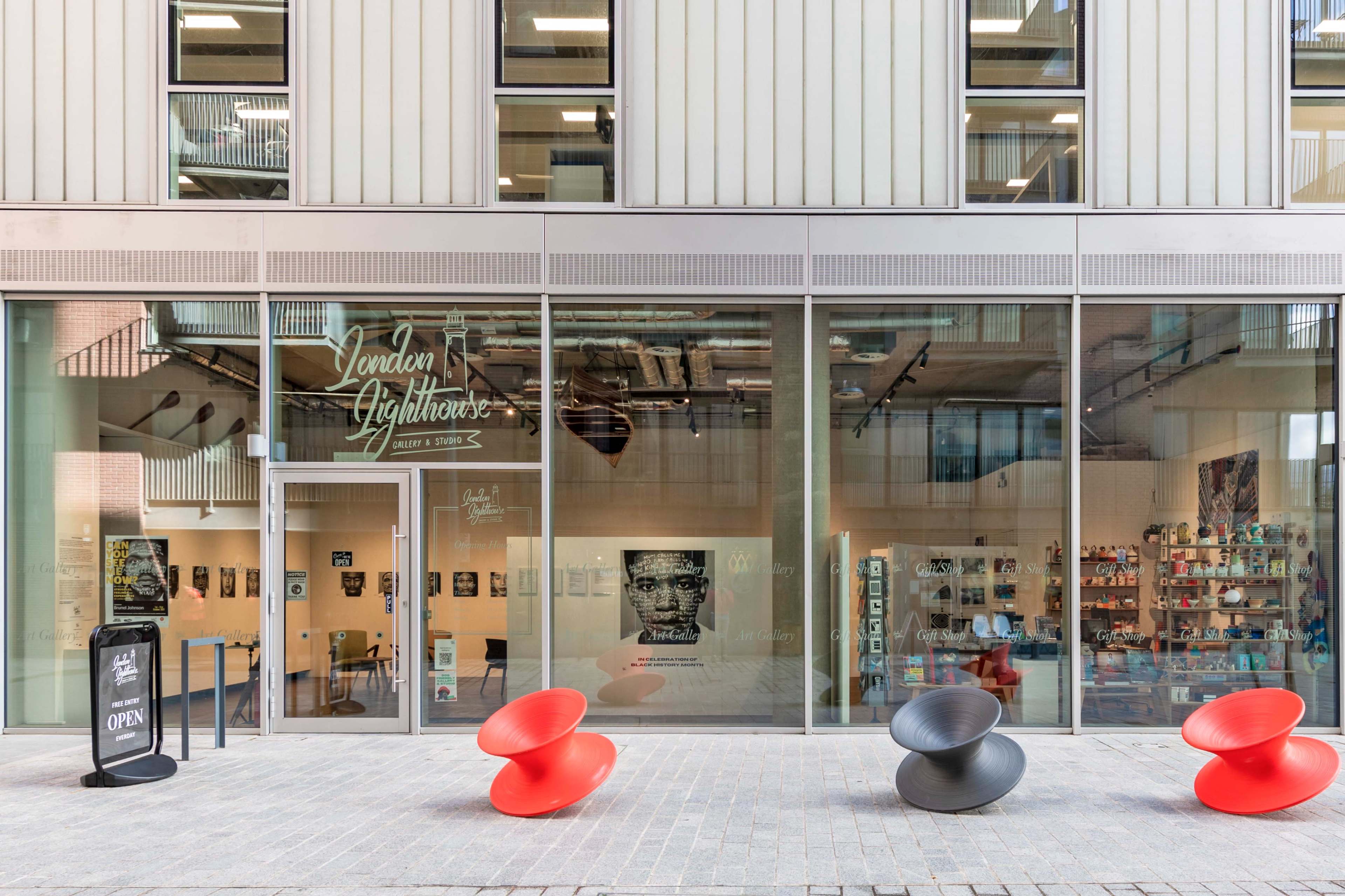 The exterior of a building with large glass windows displaying an art gallery called "London Lighthouse," featuring two colorful chairs in front.