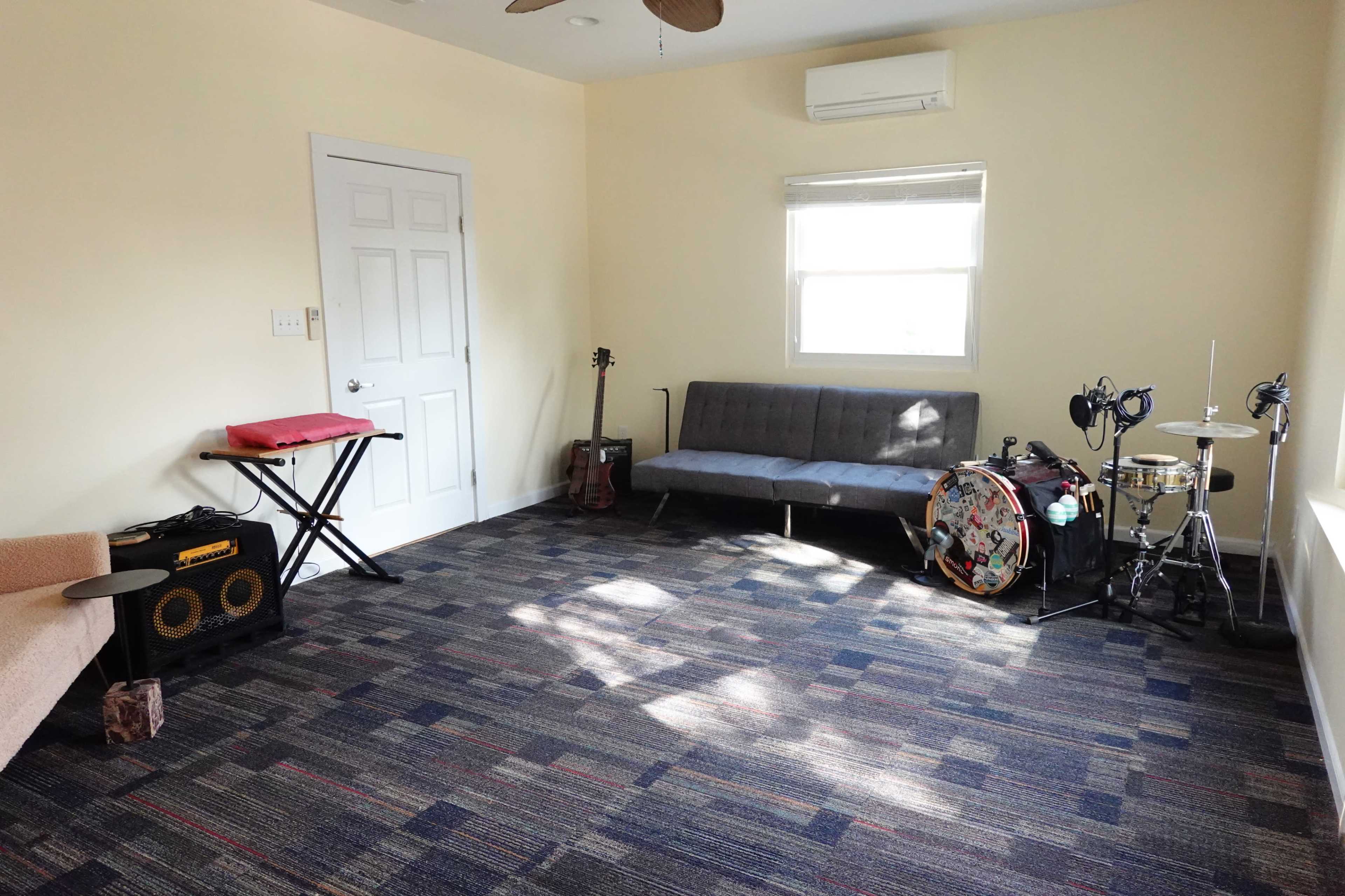 The room features a futon, a keyboard, a guitar, and a drum set, with a window letting in natural light.