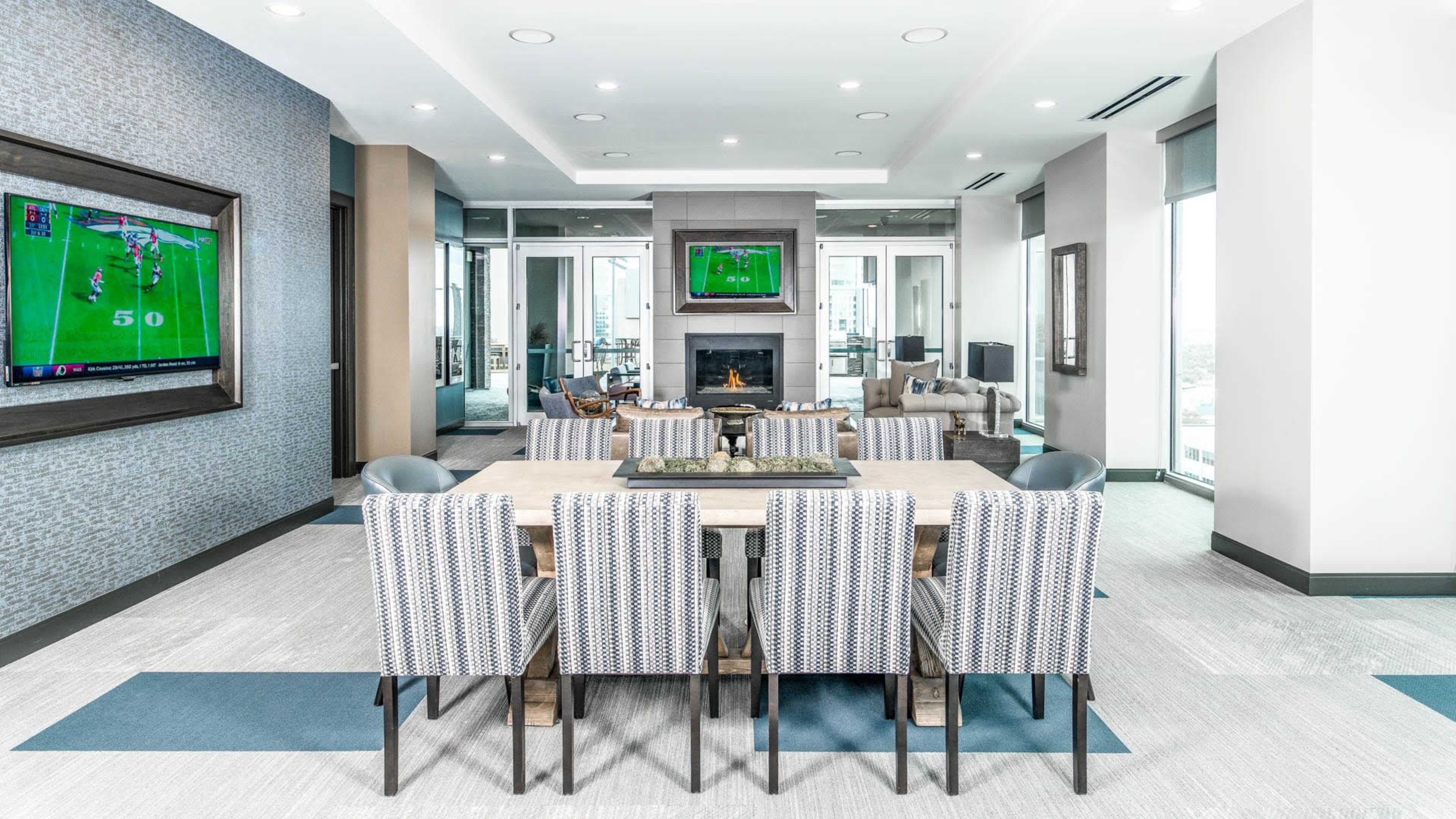 A modern lounge area with a long dining table surrounded by patterned chairs, two wall-mounted televisions, and a fireplace, all illuminated by overhead lighting.